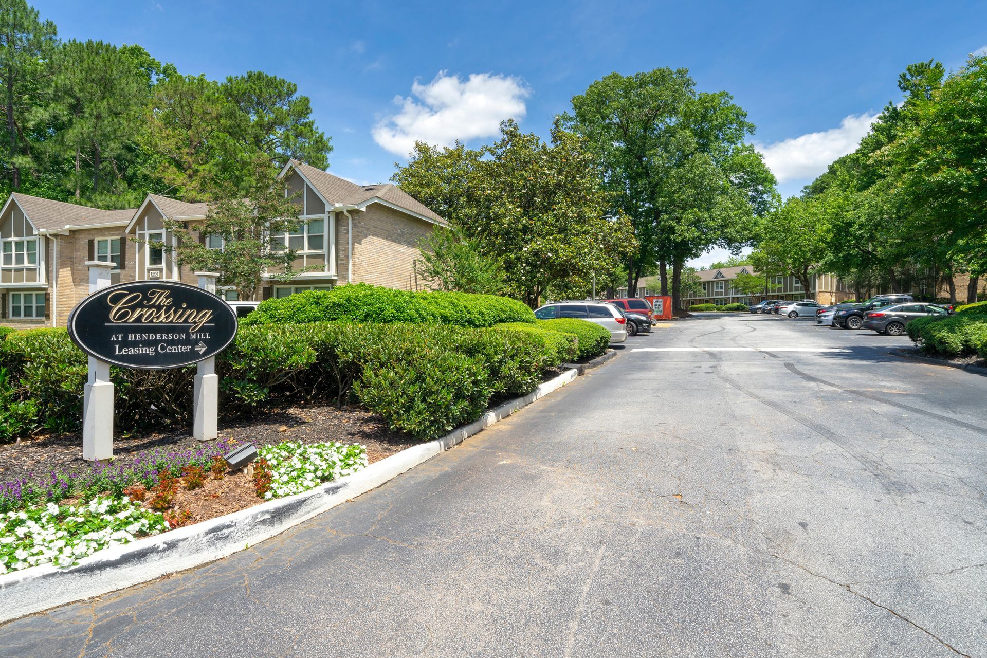 Apartment complex entrance with sign, landscaped shrubs, parked cars, and a tree-lined driveway on a sunny day