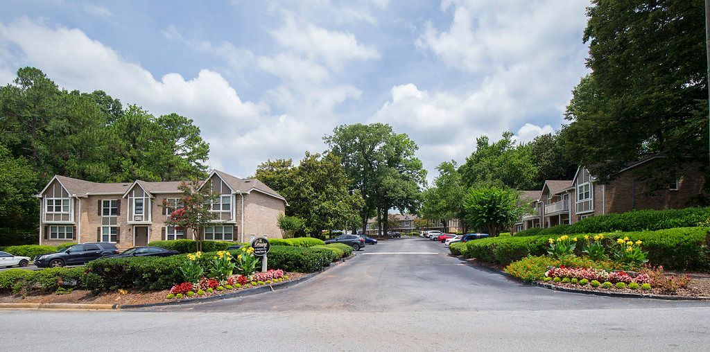 Tree-lined residential street with apartment buildings, flower beds, and a curved road under a cloudy sky