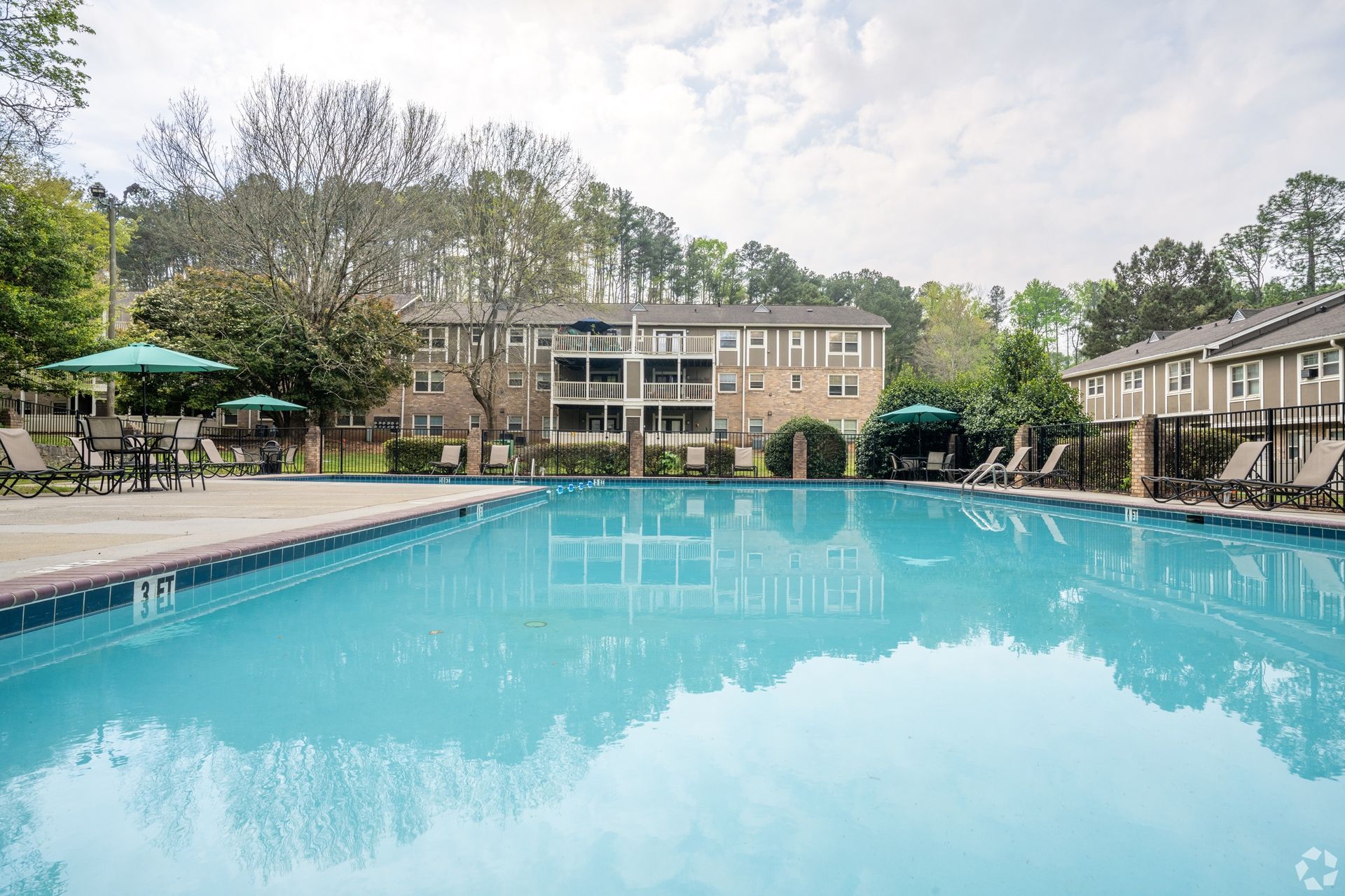 Outdoor swimming pool beside a two-story apartment building with trees and lounge chairs in the background