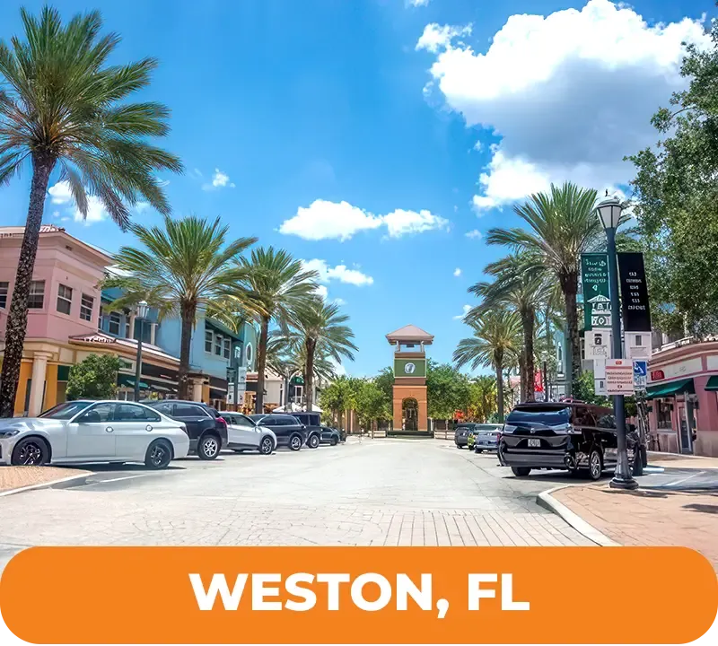 Street view in Weston, Florida with palm trees, buildings, clock tower, parked cars, and blue sky.