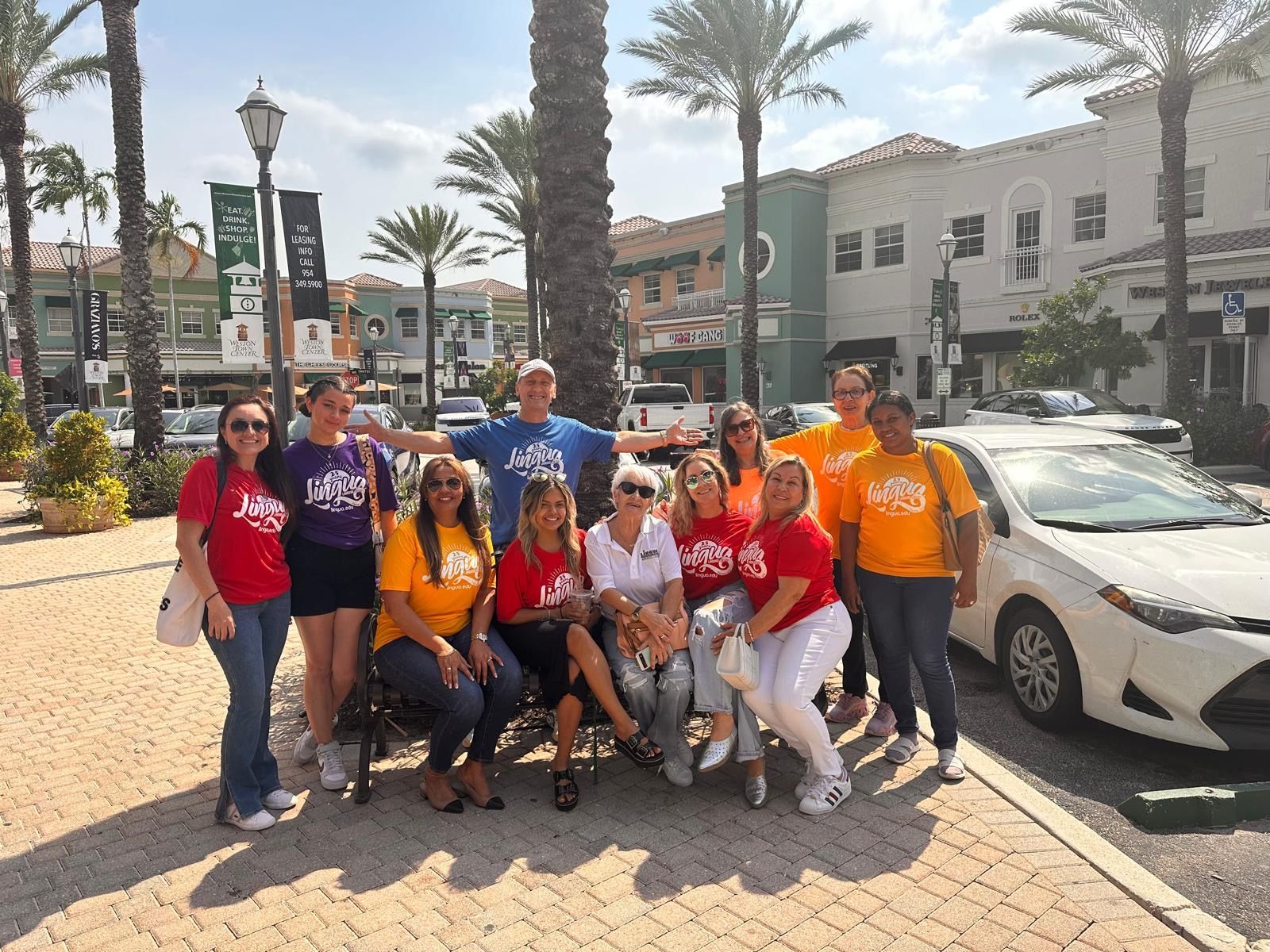 Group posing outdoors on a sunny palm-lined street beside a white car and colorful buildings.