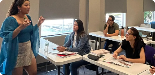 Presenter speaking to seated students in a classroom, with desks, notebooks, and bright window light.