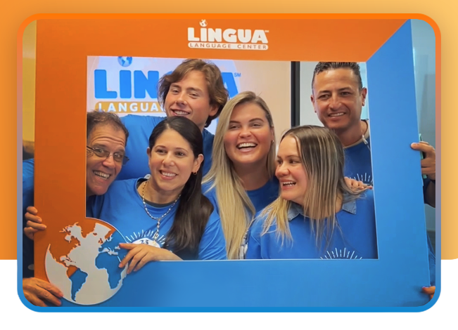 Five smiling people in blue shirts posing inside a blue photo frame at a Lingua Language School event