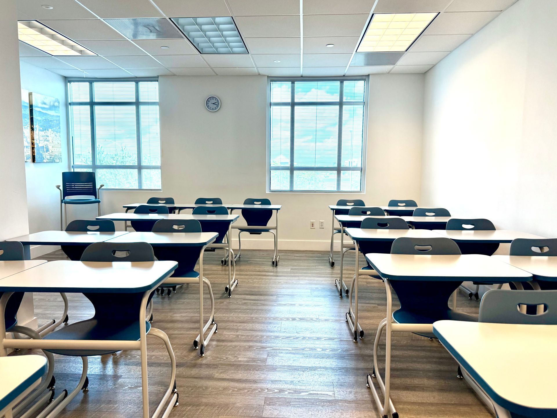 Empty classroom with rows of desks, white walls, and large windows letting in daylight.