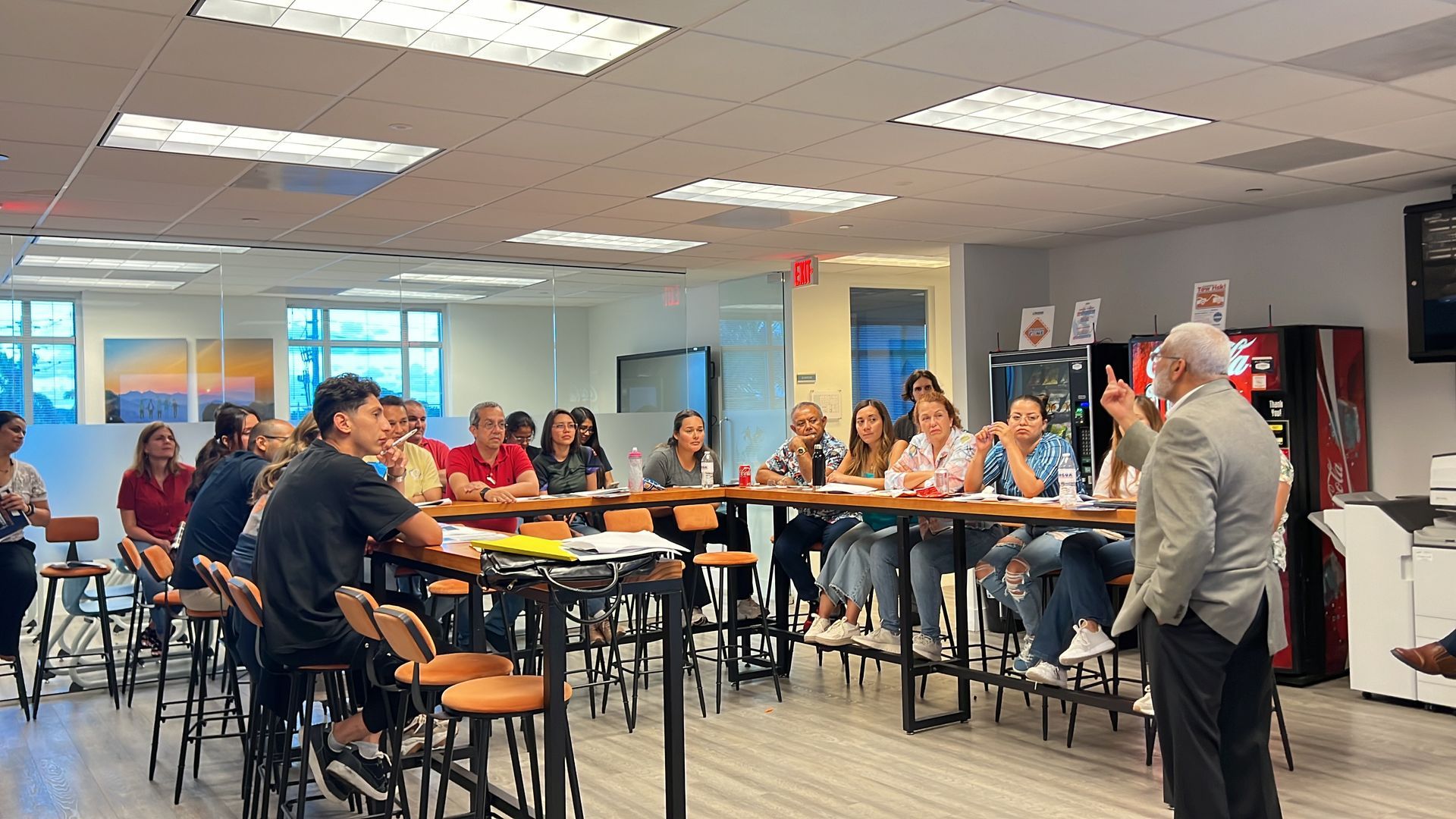 Meeting in a conference room with people seated around tables listening to a speaker at the front