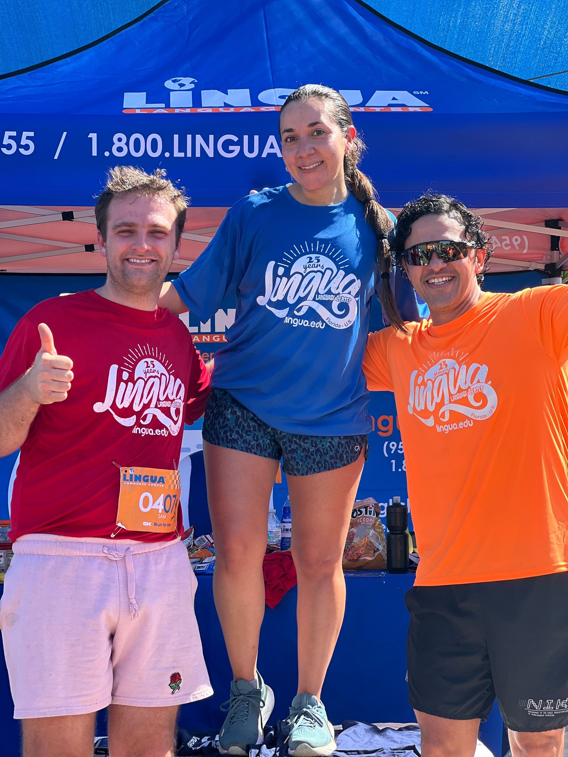 Three people posing with thumbs up in front of a blue event tent, wearing matching T-shirts
