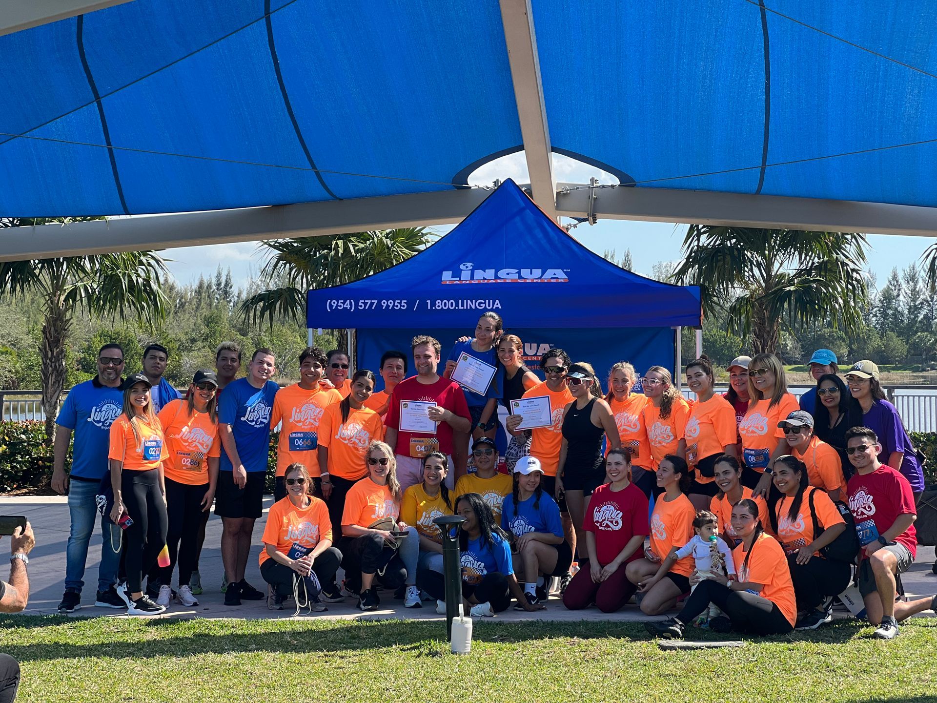 Group of people in orange and blue shirts posing under a blue canopy at an outdoor event