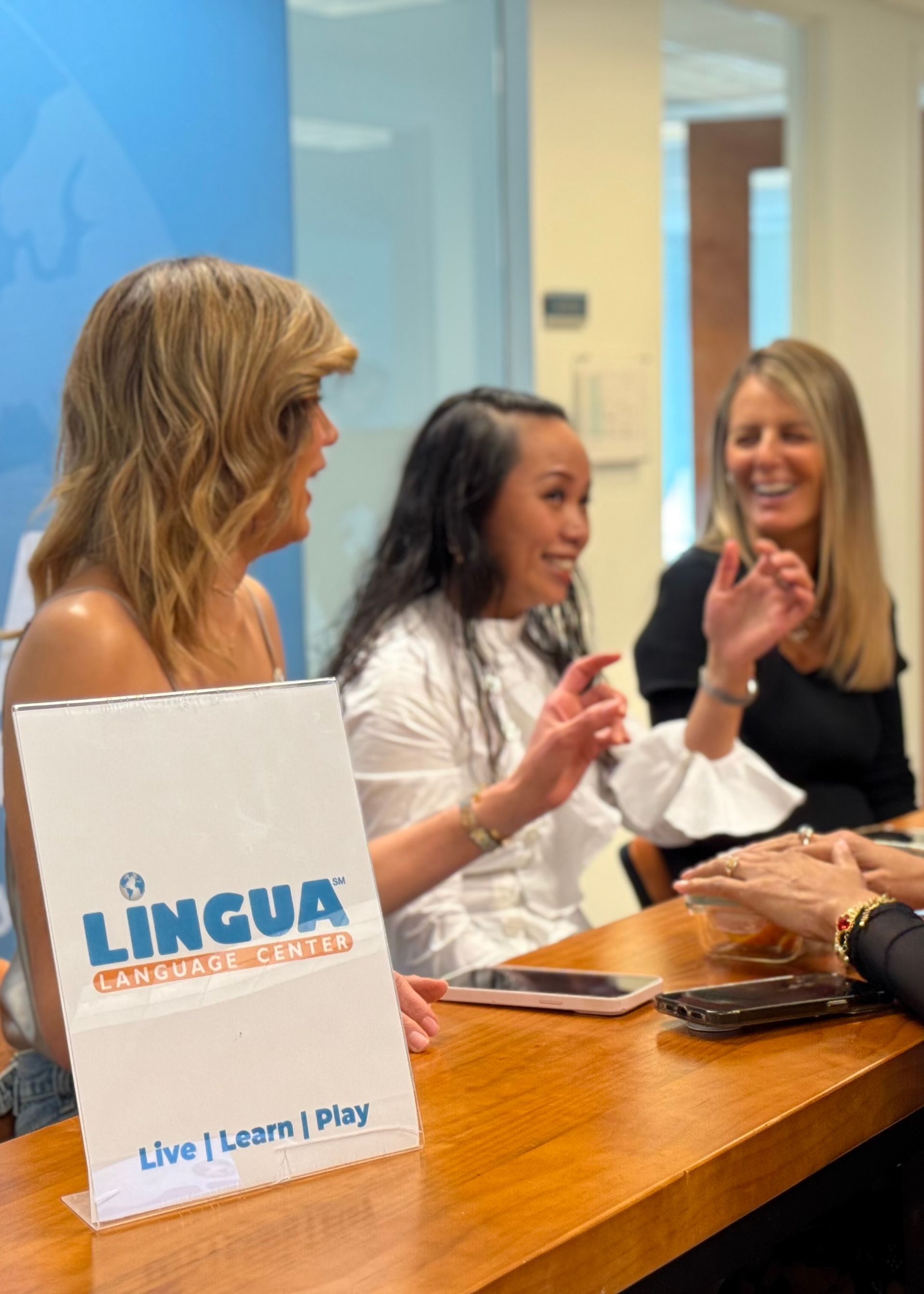 Three women chatting at a table with a Lingua sign in a bright office setting.