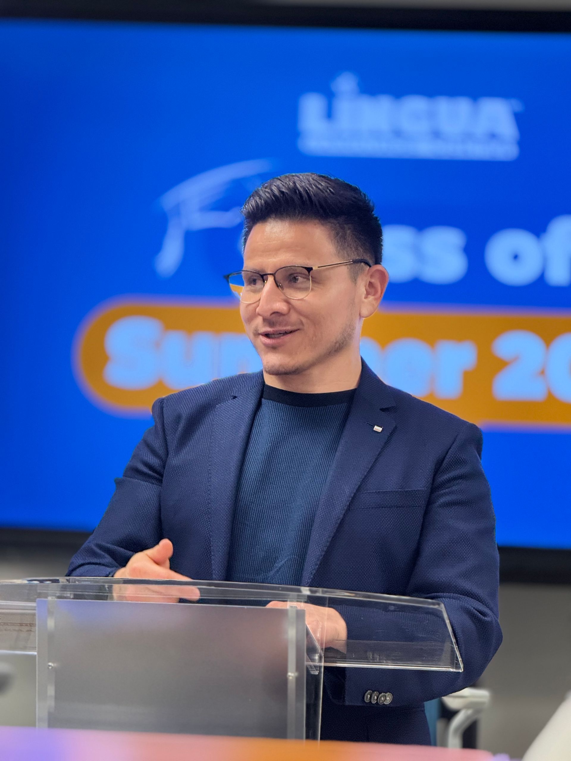 Man speaking at a podium on a stage with a blue backdrop and event signage