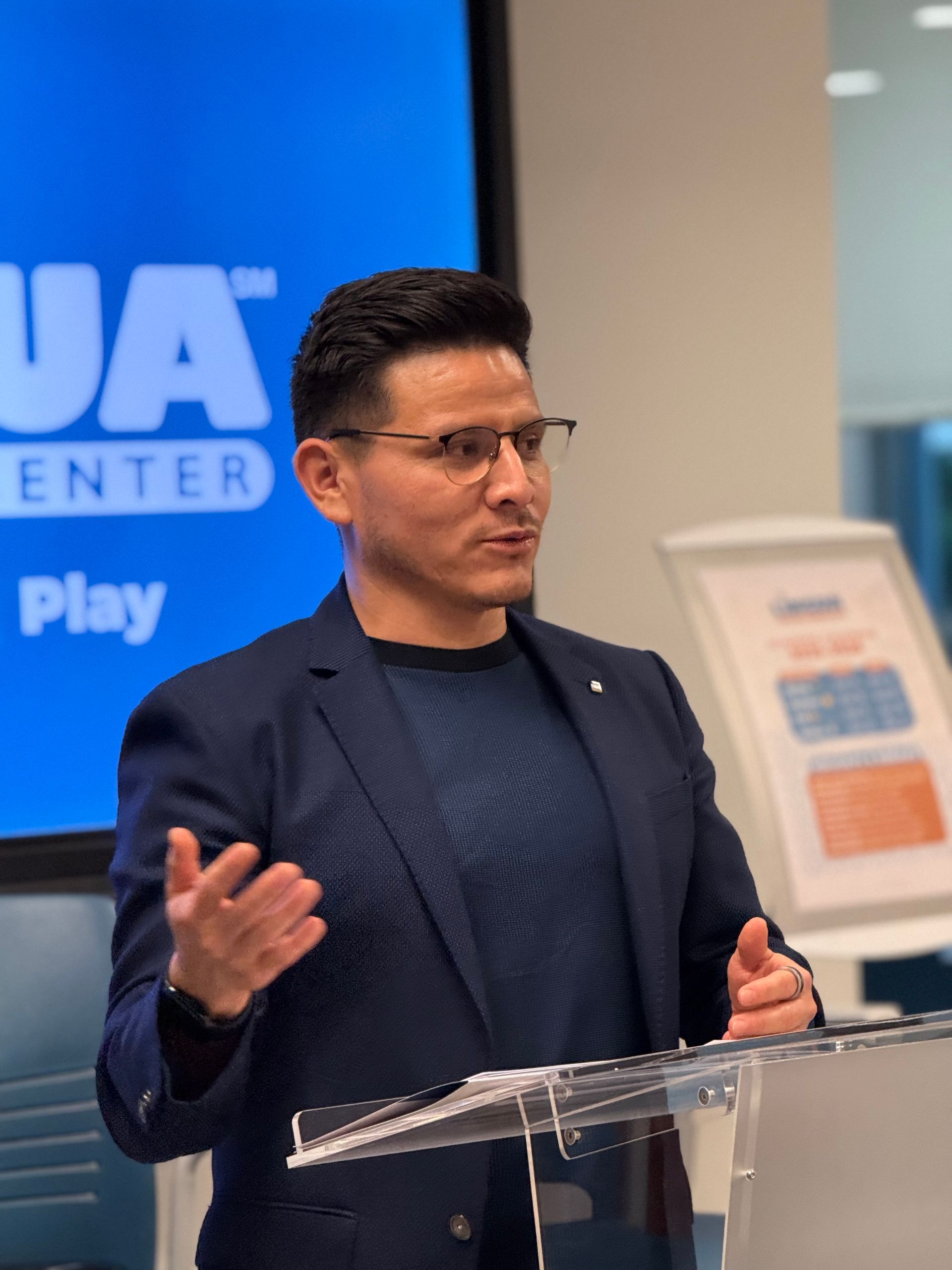 Speaker in a navy blazer gestures at a podium during a presentation, with a blue screen in the background.