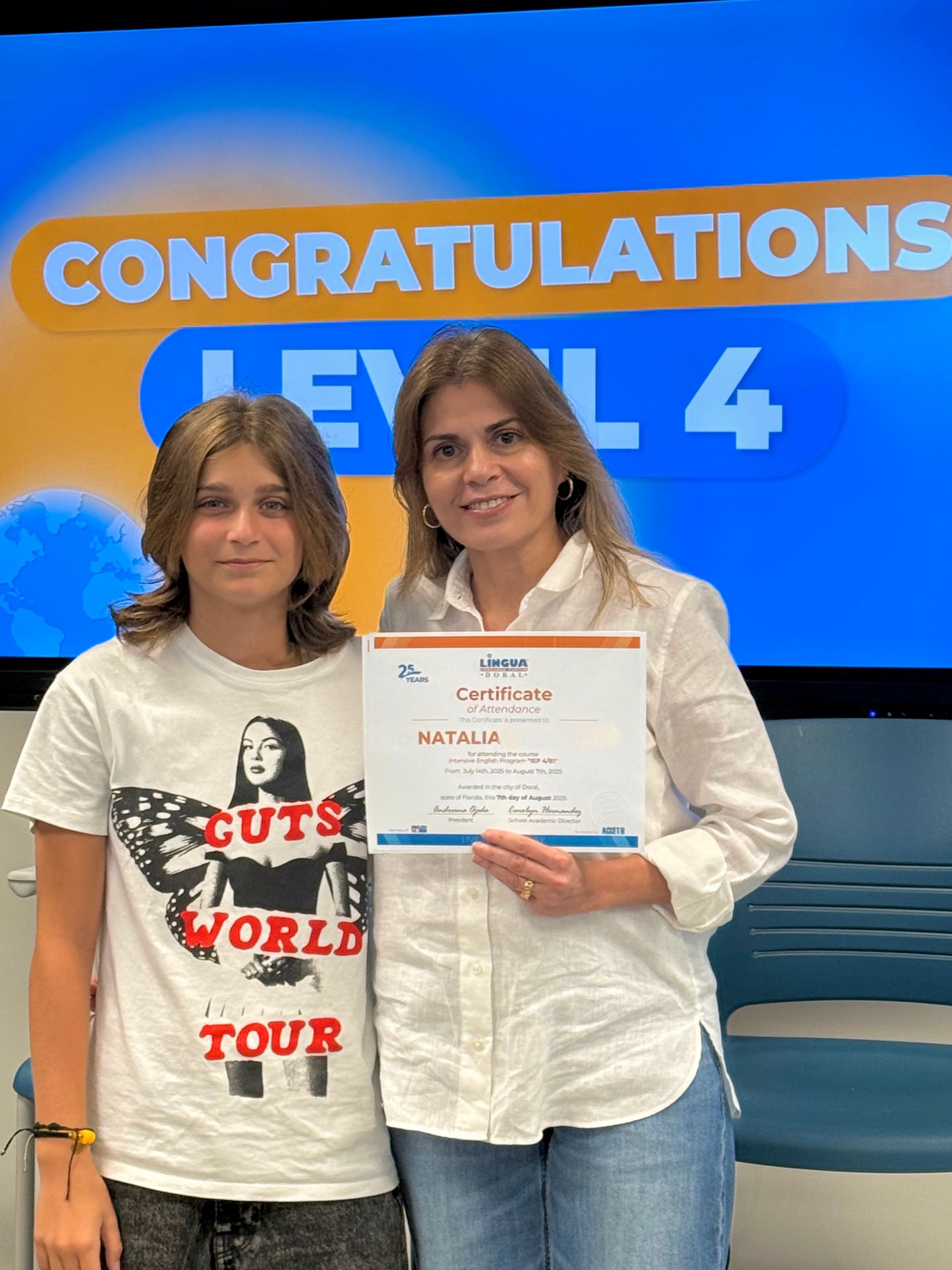Two people pose with a certificate in front of a blue “Congratulations Level 4” backdrop.