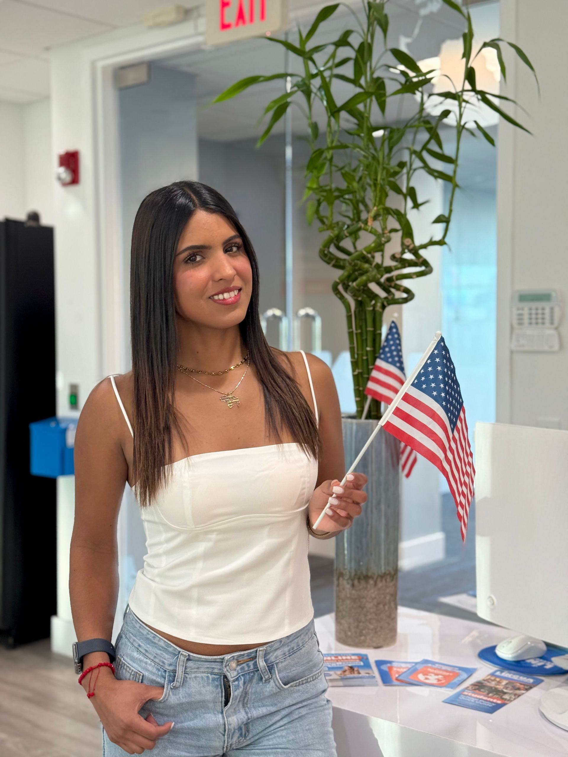 Woman holding an American flag in a bright indoor hallway, smiling beside a table with patriotic materials.