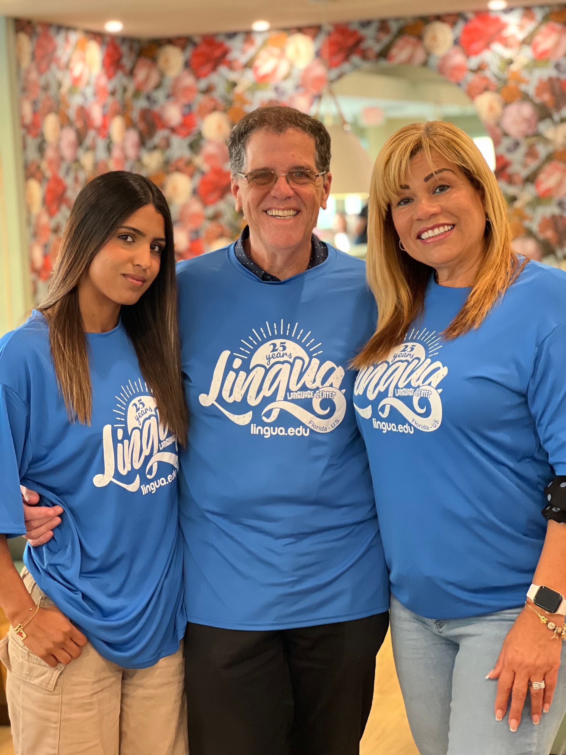Three people in blue logo T-shirts smiling in front of a floral backdrop.