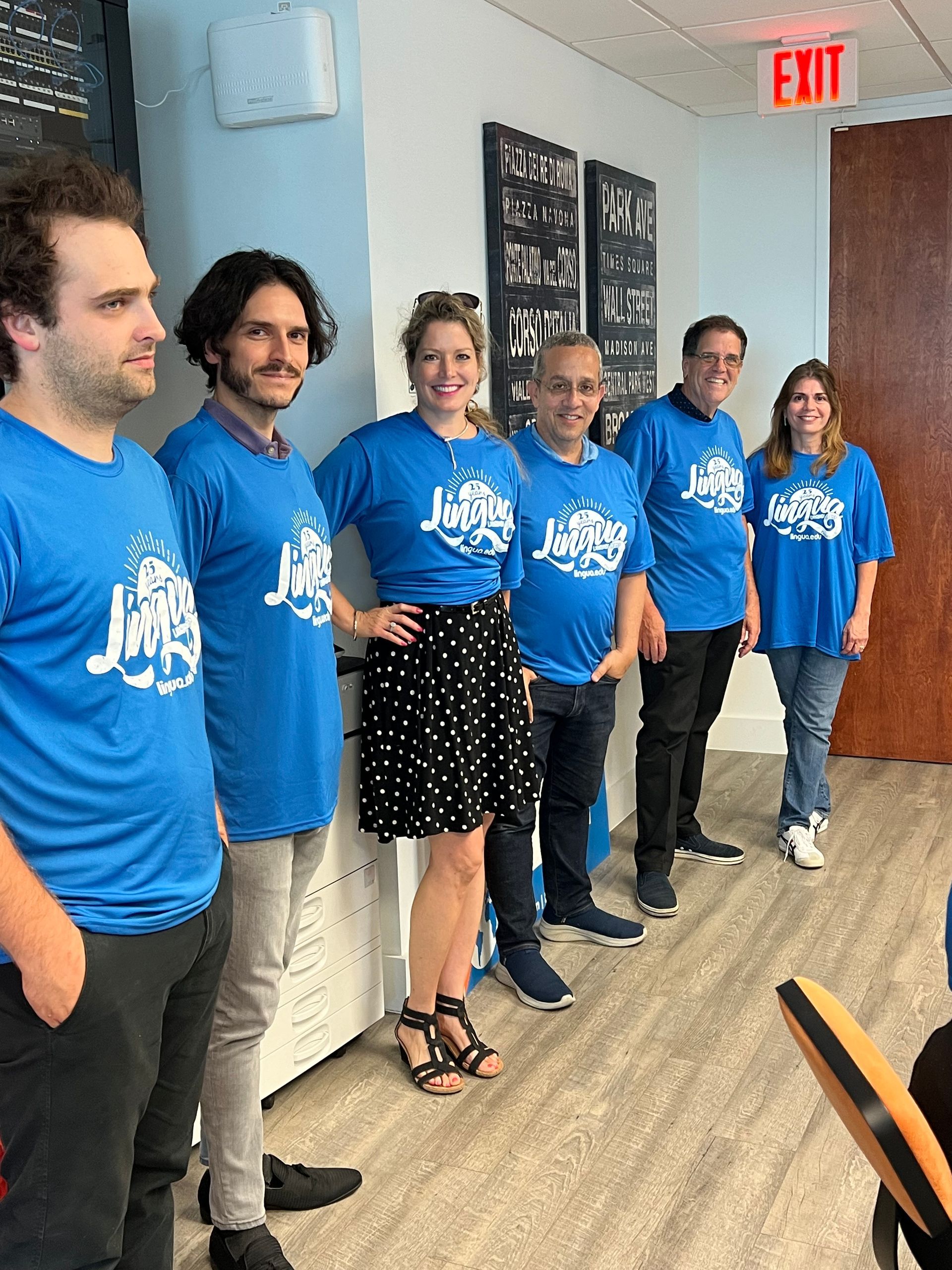 People in blue event shirts line a hallway beside a table, smiling for a group photo.