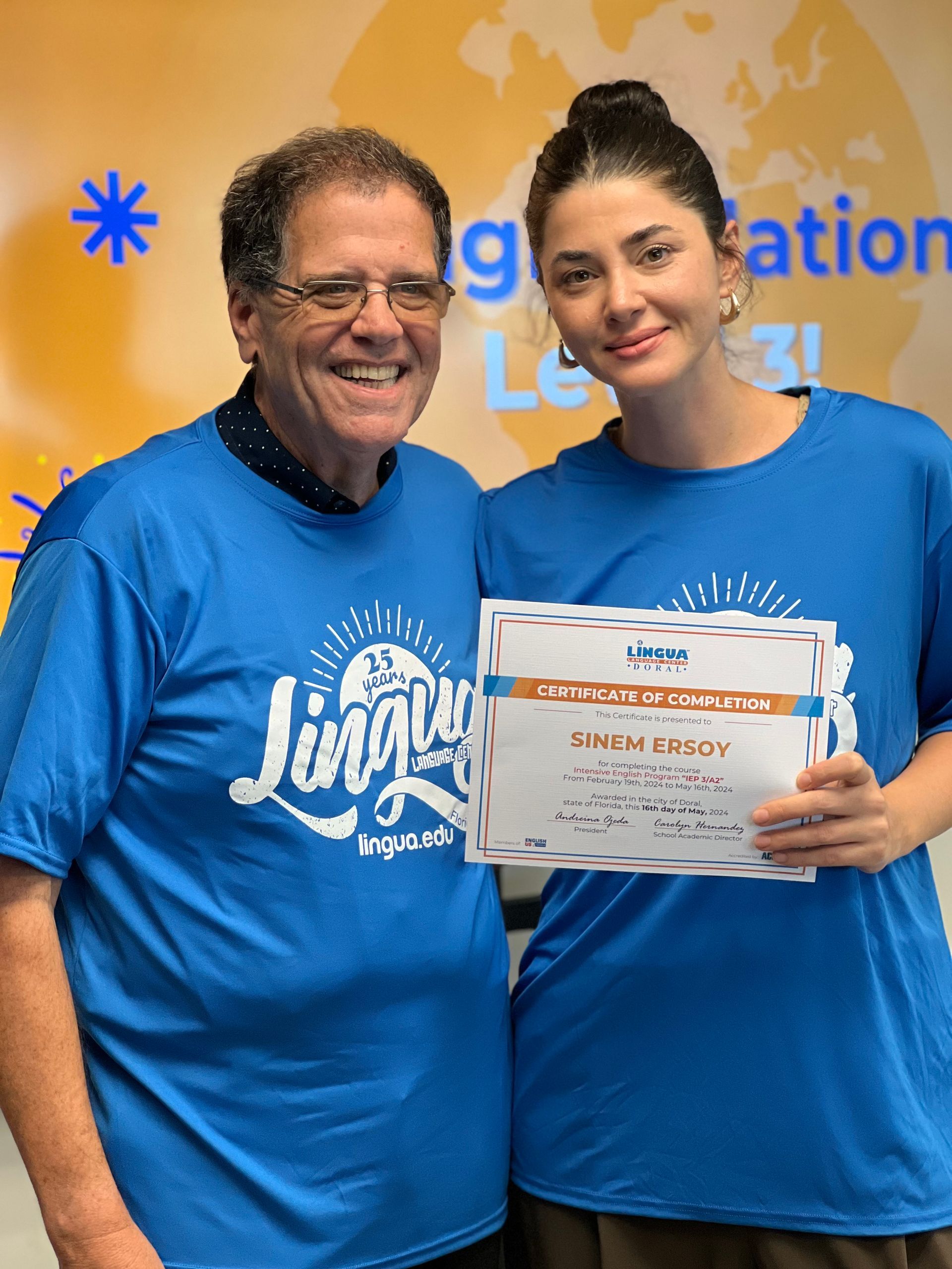 Two people in blue shirts holding a certificate and smiling in front of a graduation backdrop