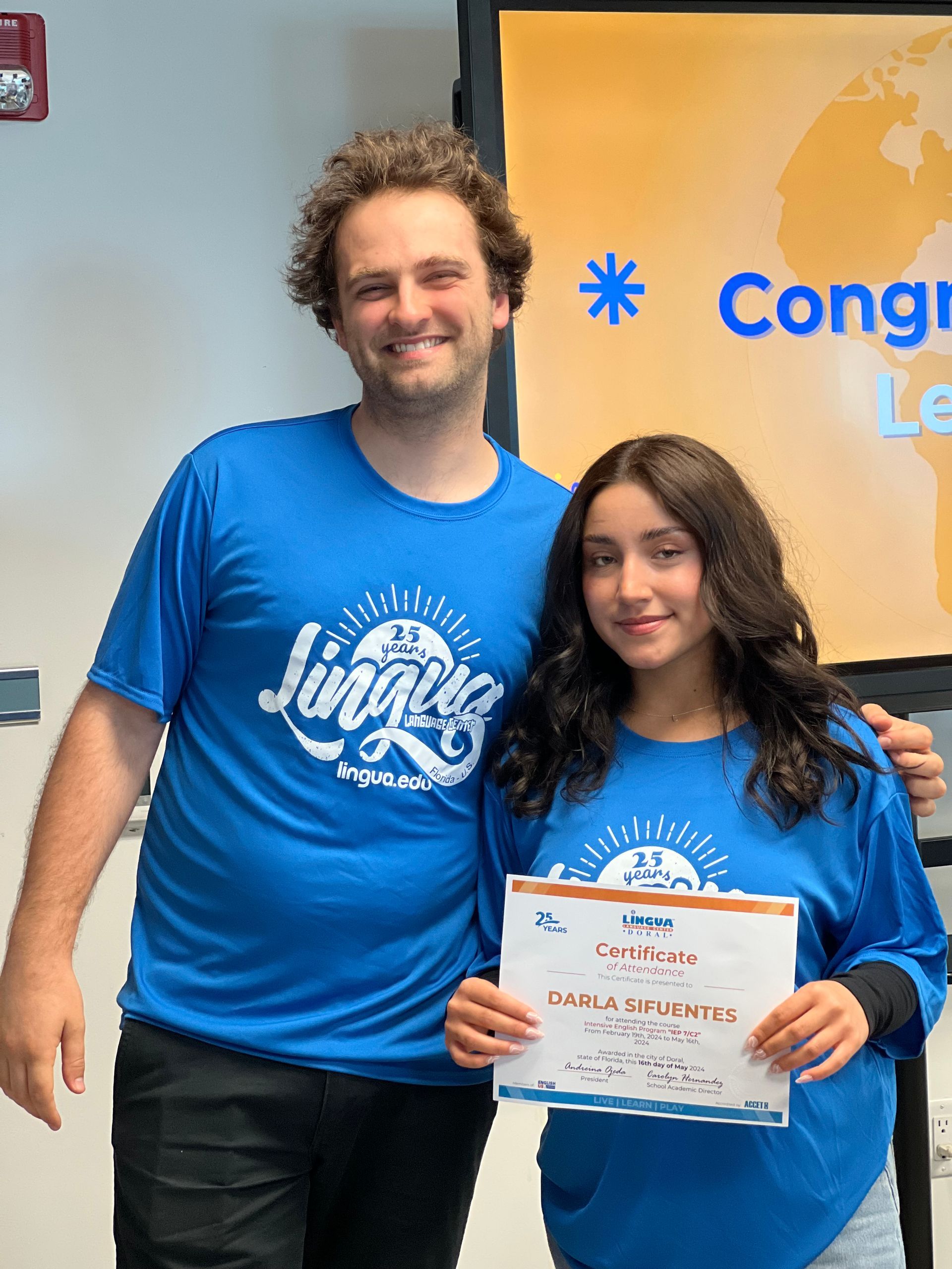 Two people in blue shirts pose indoors, smiling and holding a certificate.