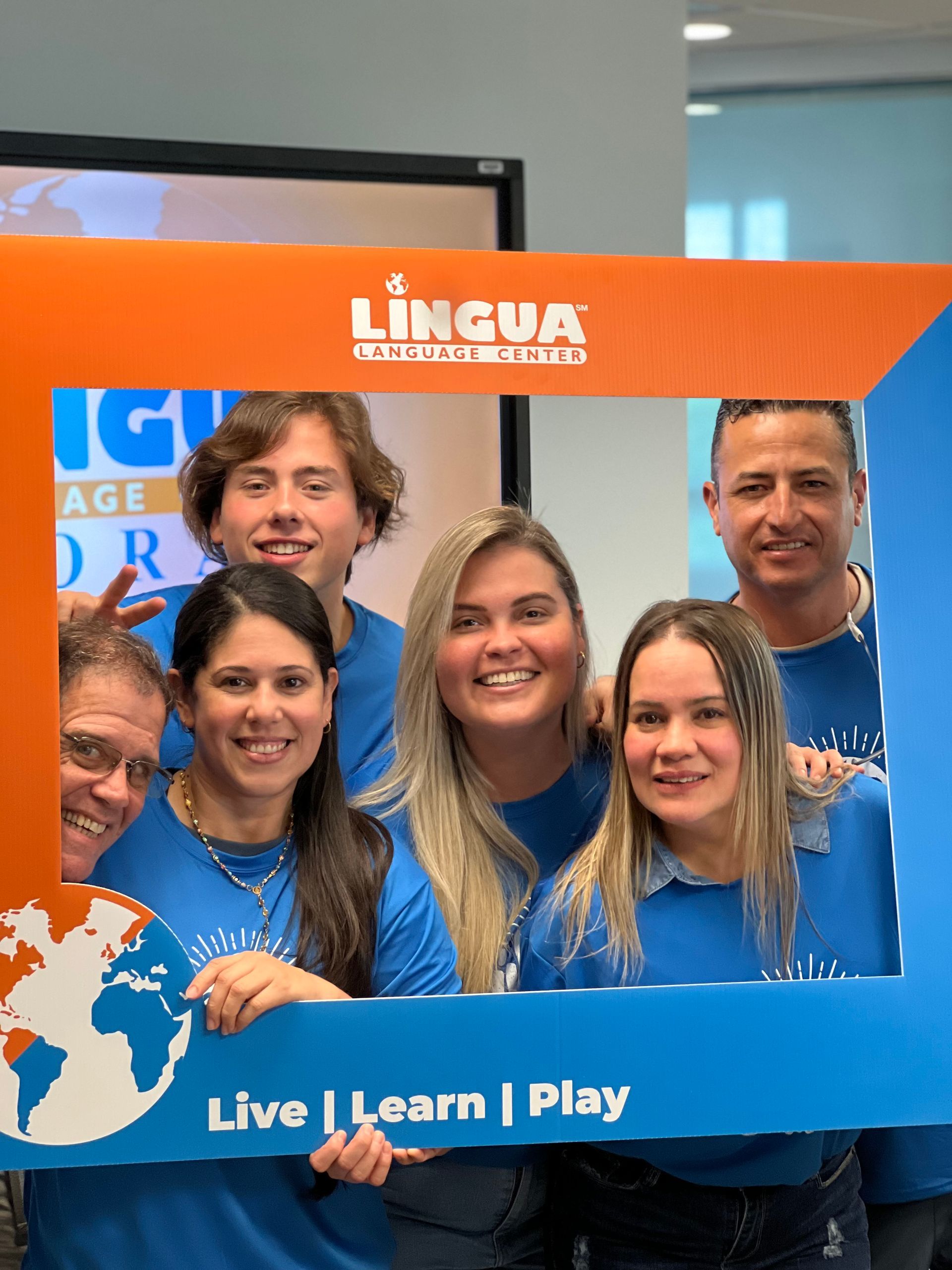 Five smiling people posing behind a blue and orange LINGUA banner with “Live | Learn | Play” text.