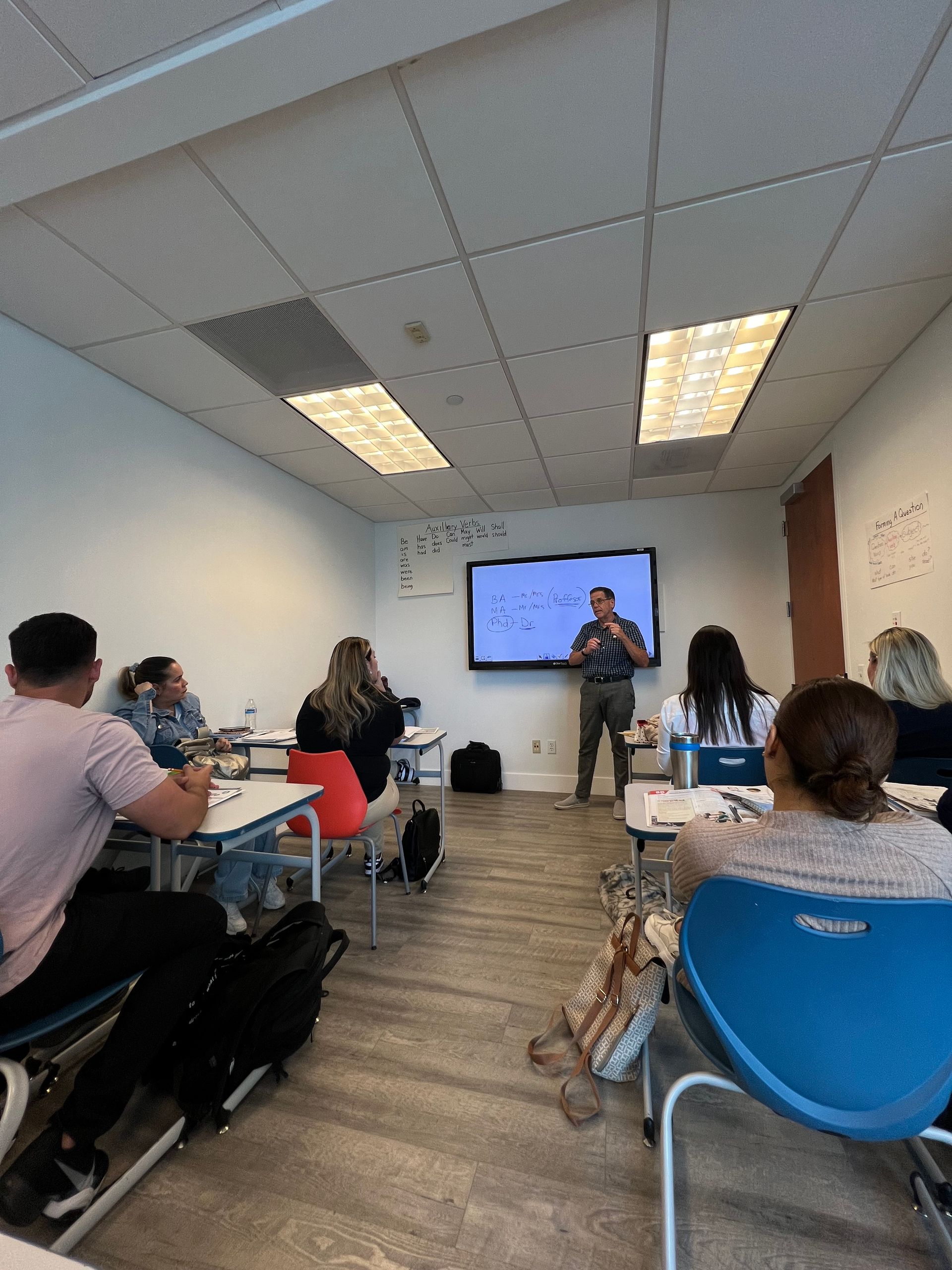 Small classroom presentation with people seated at desks facing a speaker near a wall-mounted screen.