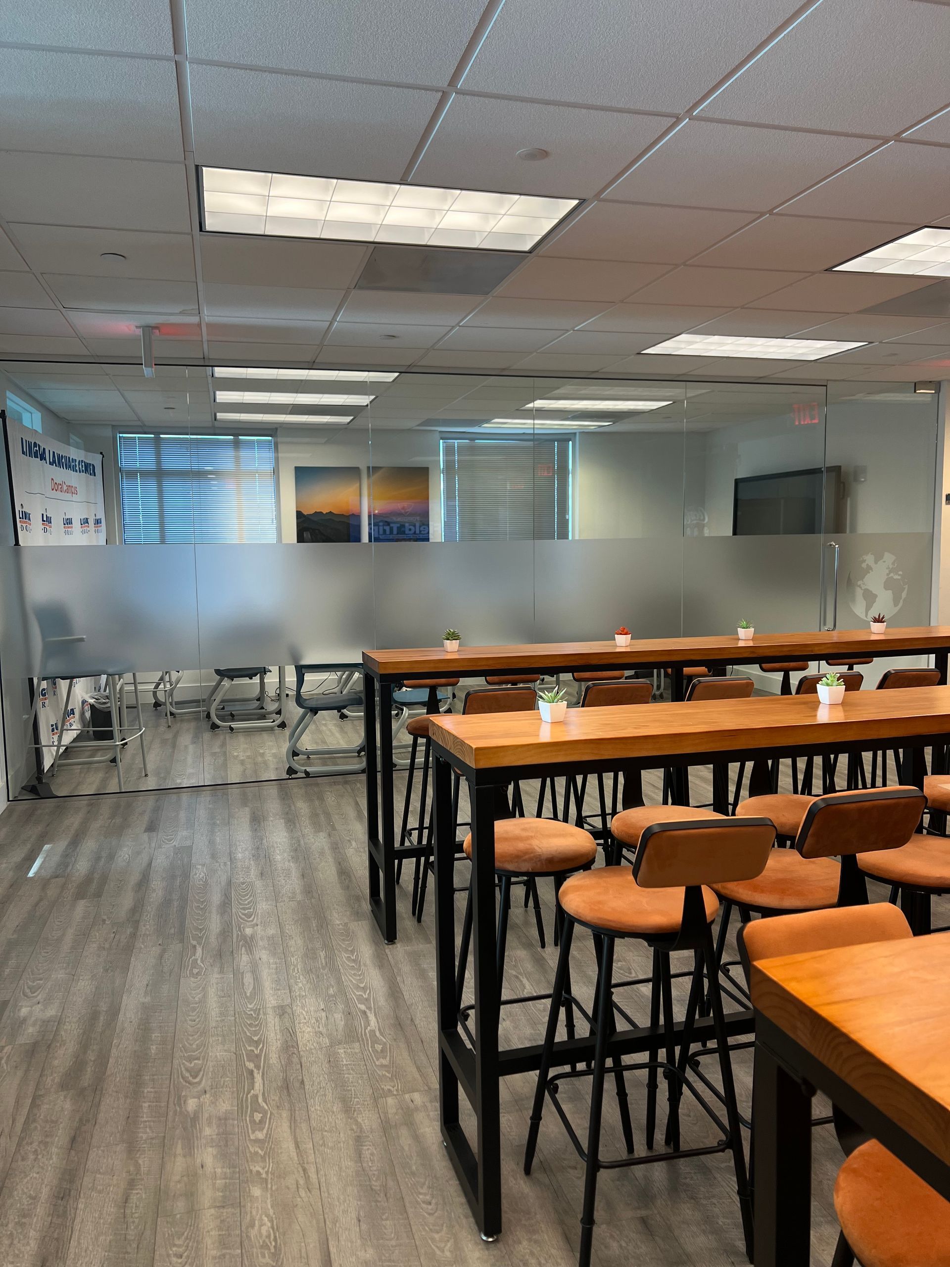 Empty classroom with rows of wooden tables and stools, frosted glass wall, and ceiling lights.