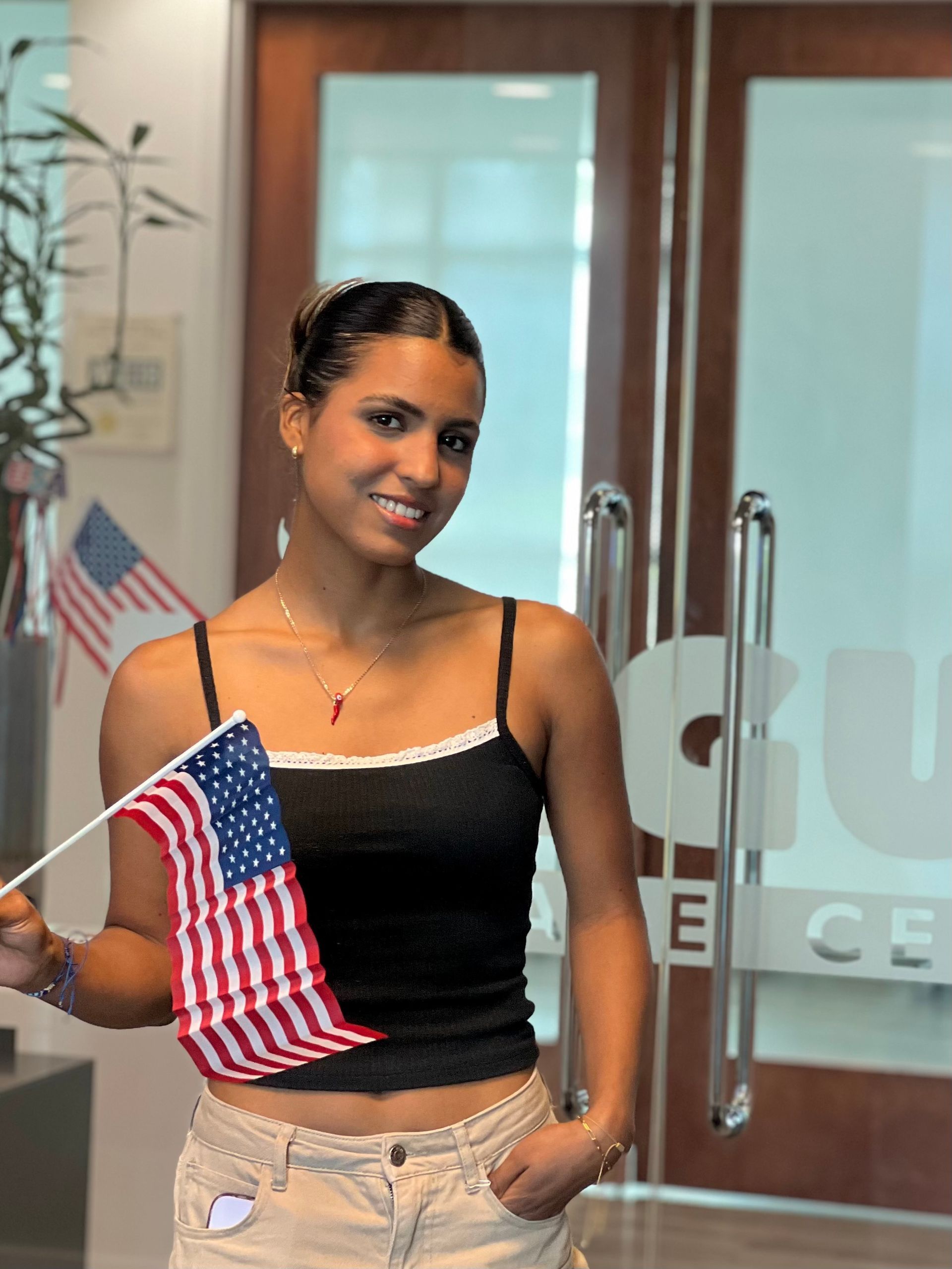 Woman holding a small American flag in front of glass doors, wearing a black top and beige pants.
