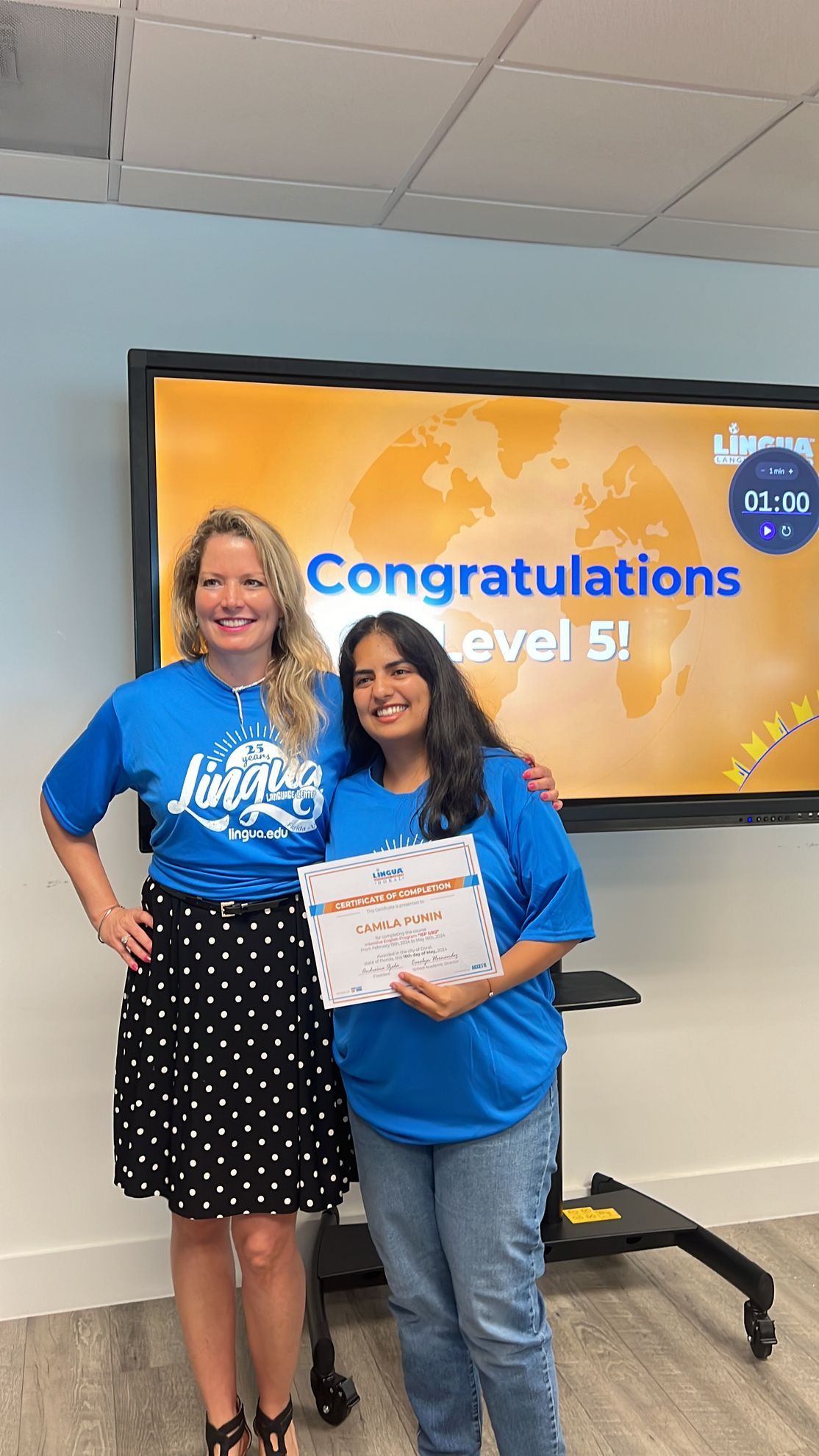 Two women in blue shirts pose with a certificate in front of a “Congratulations Level 5” screen.