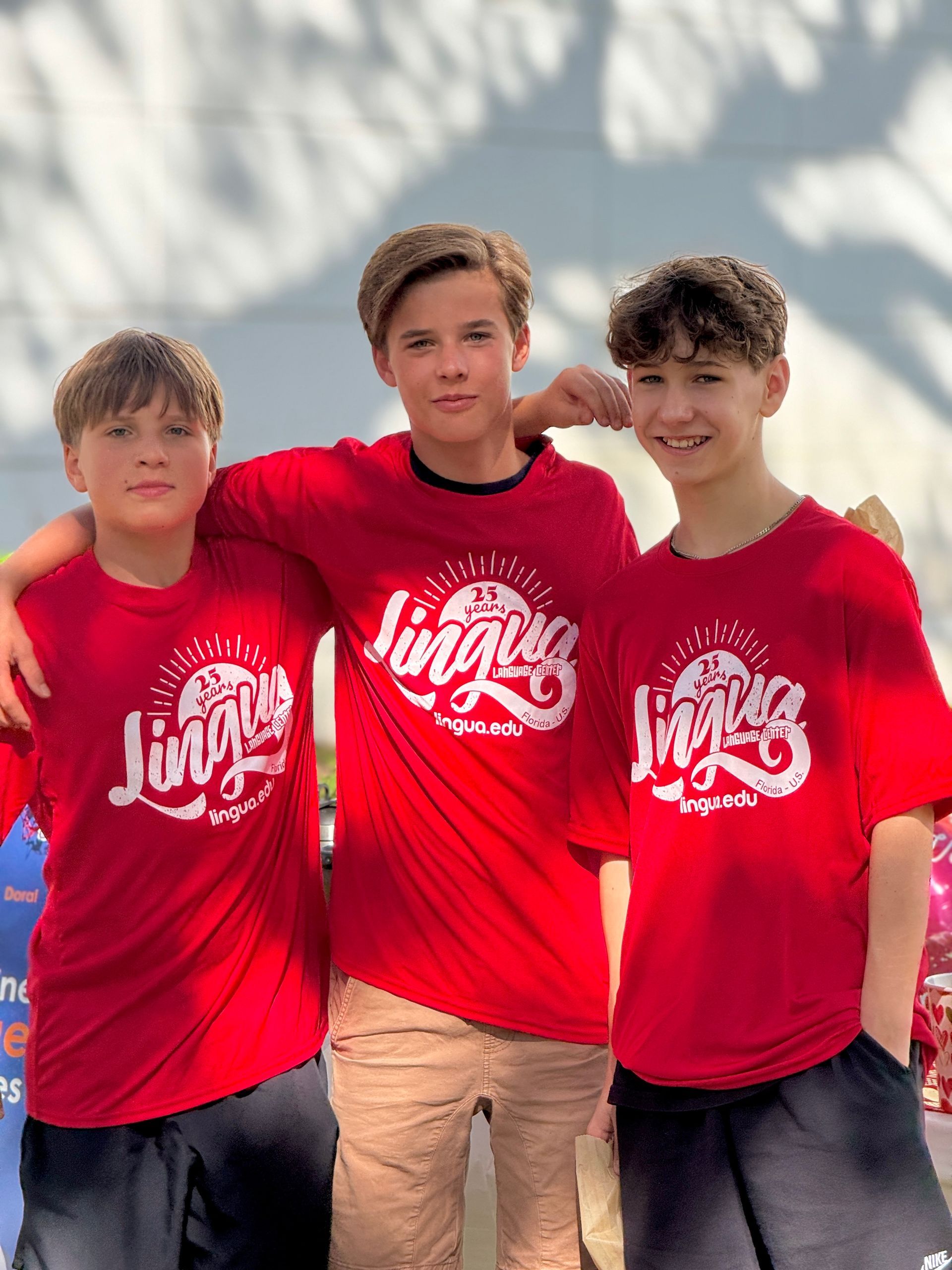 Three friends in matching red shirts stand together outdoors, smiling at the beach.