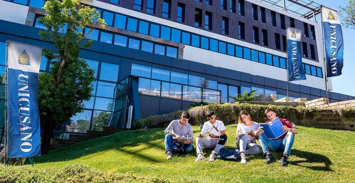 People sitting on a grassy lawn outside a blue glass office building with vertical banners.