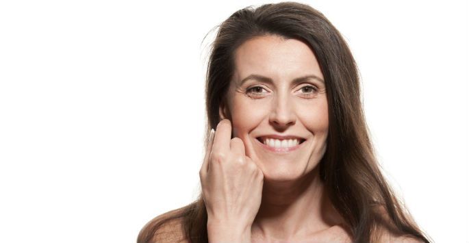 A woman with long hair is smiling and touching her face.
