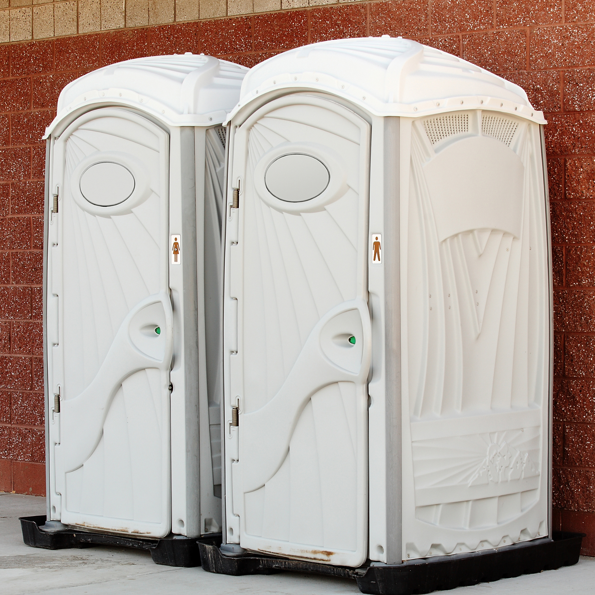 Two white portable toilets standing side by side against a red brick wall.