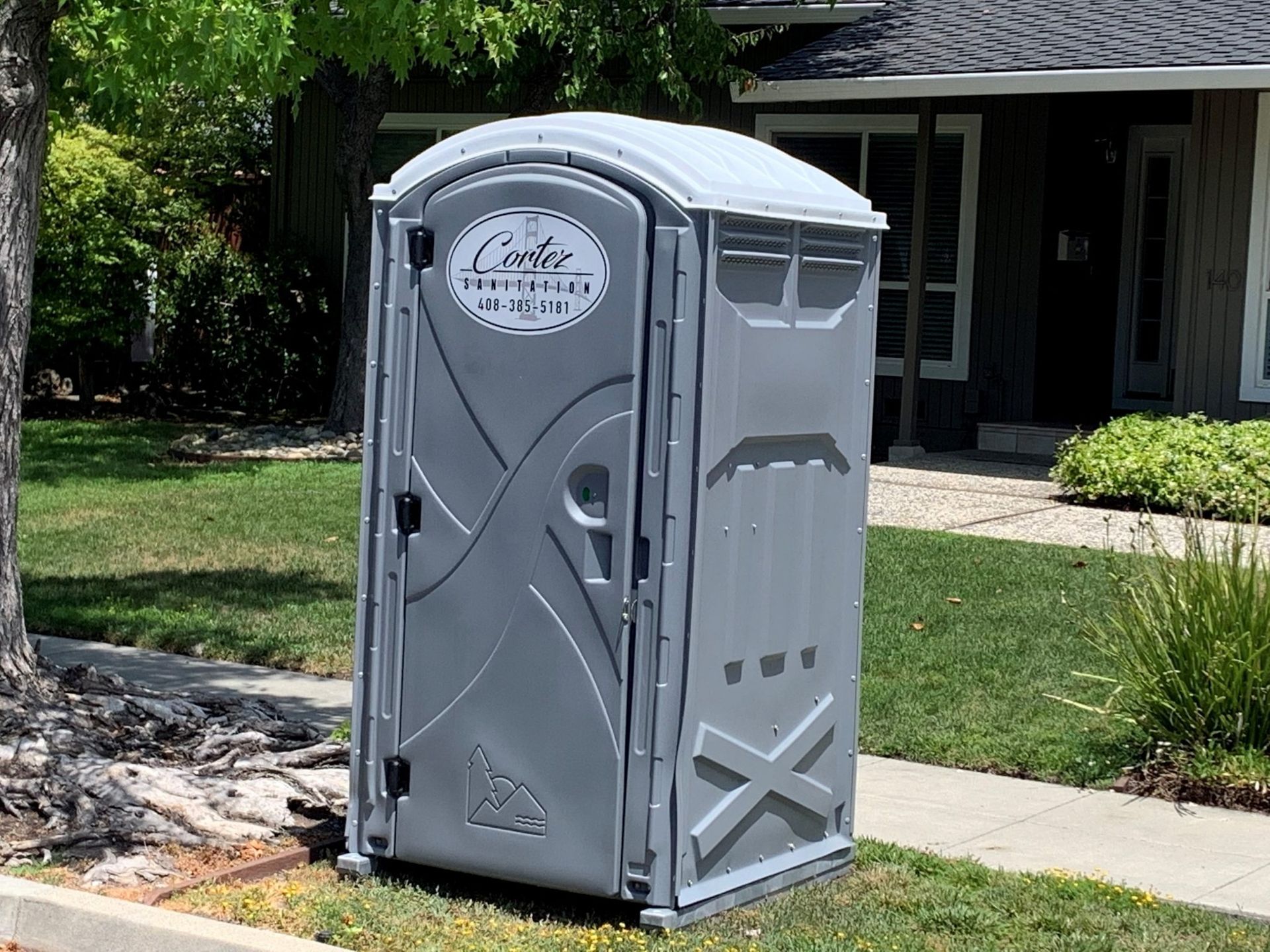 Gray portable toilet on a sidewalk beside a grassy yard and house entrance.