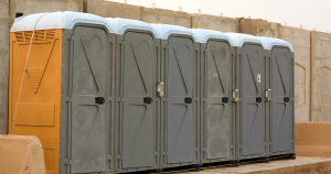 Row of gray portable toilets with an orange one at the end, set against a tan wall with snow on top.