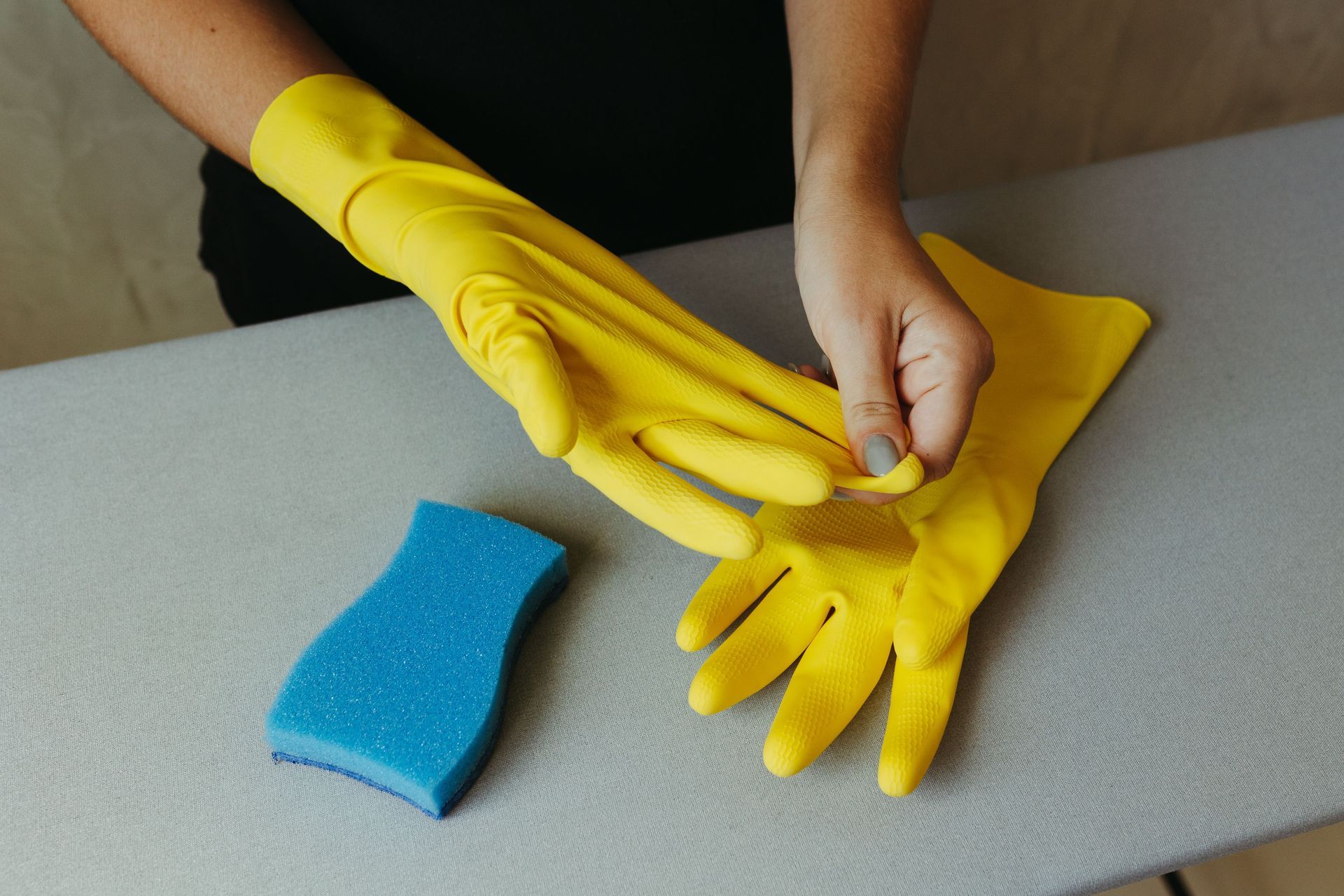 Hands putting on bright yellow rubber gloves beside a blue sponge on a table