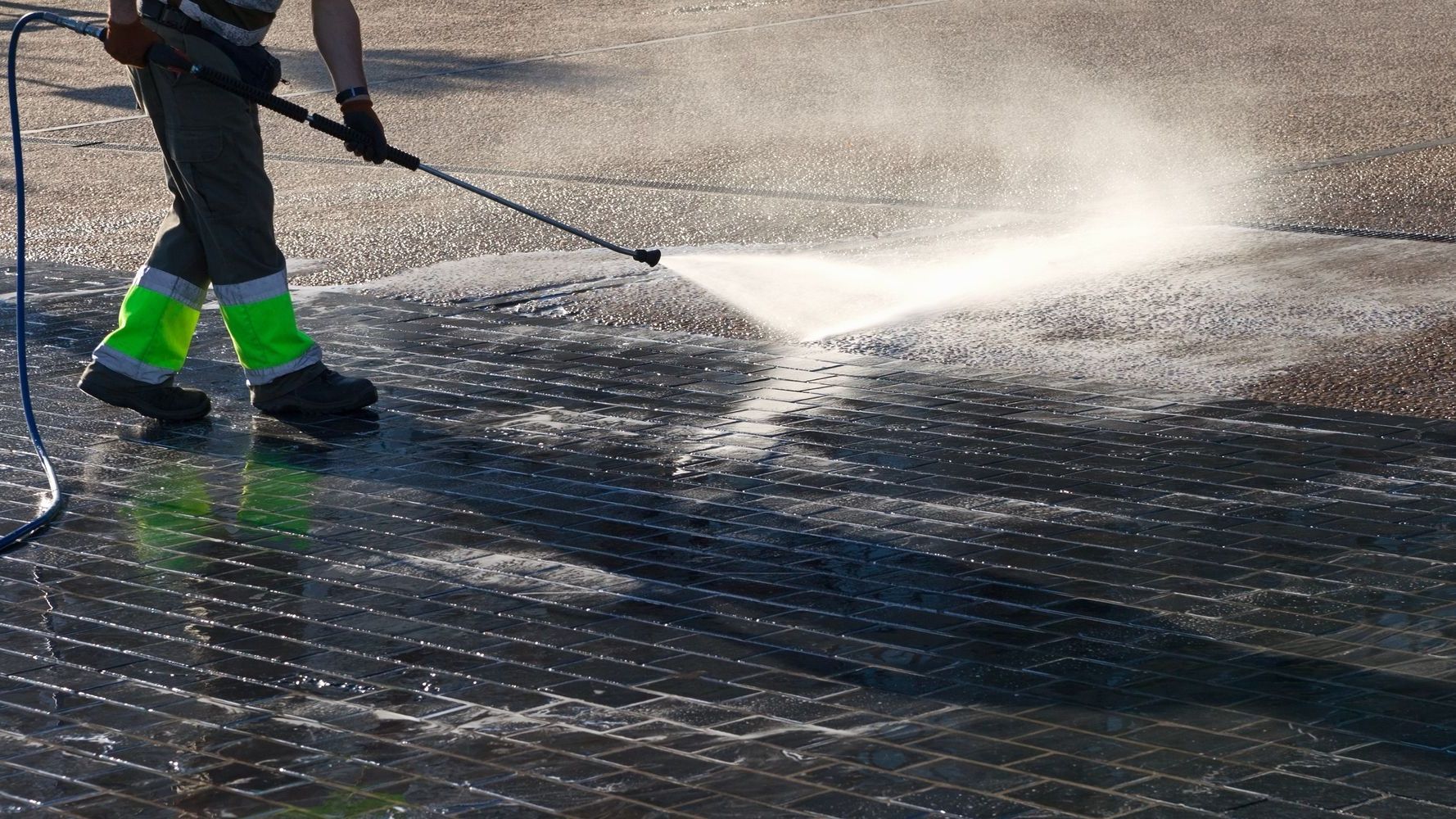 Une personne nettoie une surface sombre avec un jet haute pression. L'eau gicle sur le trottoir, créant une zone humide.