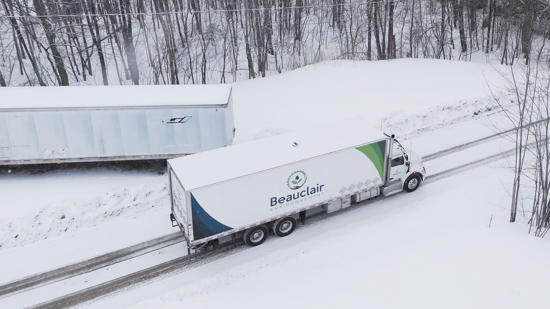 Un camion avec une remorque blanche, portant le logo « Beaulieu », roule sur une route enneigée. Une autre remorque est garée à proximité.