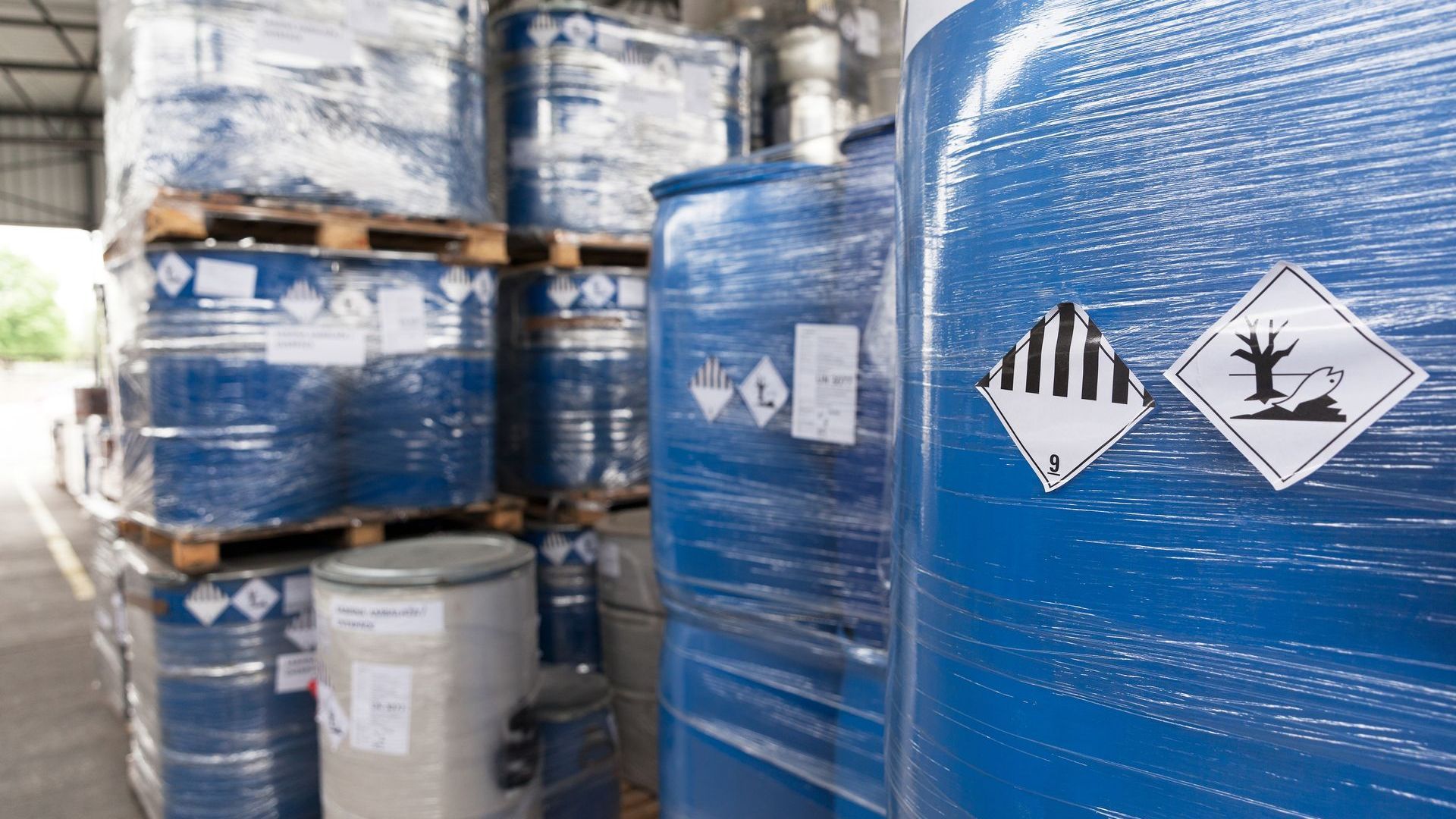 Des barils bleus de produits chimiques dangereux empilés dans un entrepôt, avec des étiquettes d'avertissement visibles.