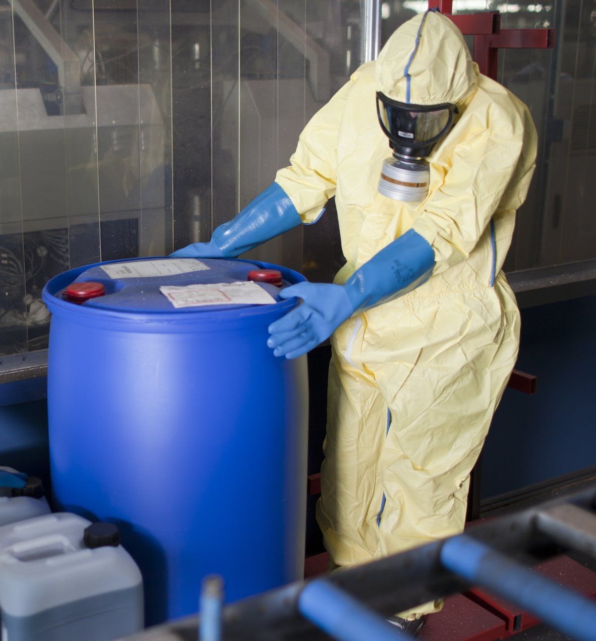 Personne portant une combinaison de protection et un respirateur à proximité d'un fût chimique bleu.