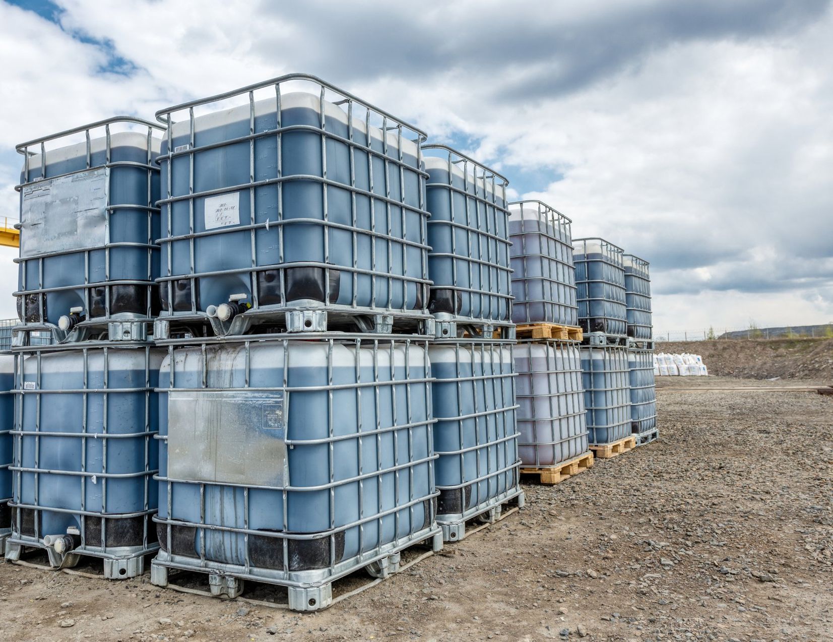 Plusieurs grands conteneurs de stockage de liquide bleu empilés sur des palettes à l'extérieur sous un ciel nuageux.