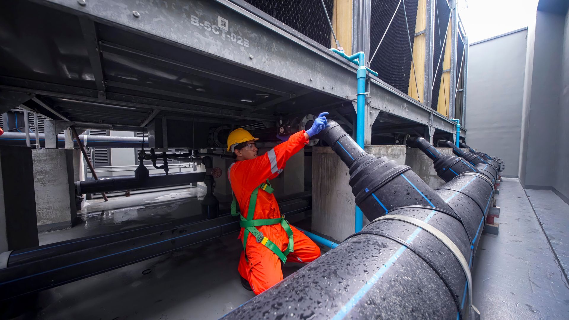 Worker in orange safety gear handling a large black pipe under industrial machinery.