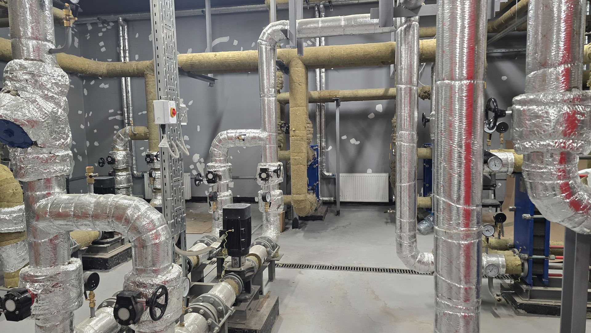 Industrial mechanical room with silver insulated pipes, valves, and equipment on a concrete floor.