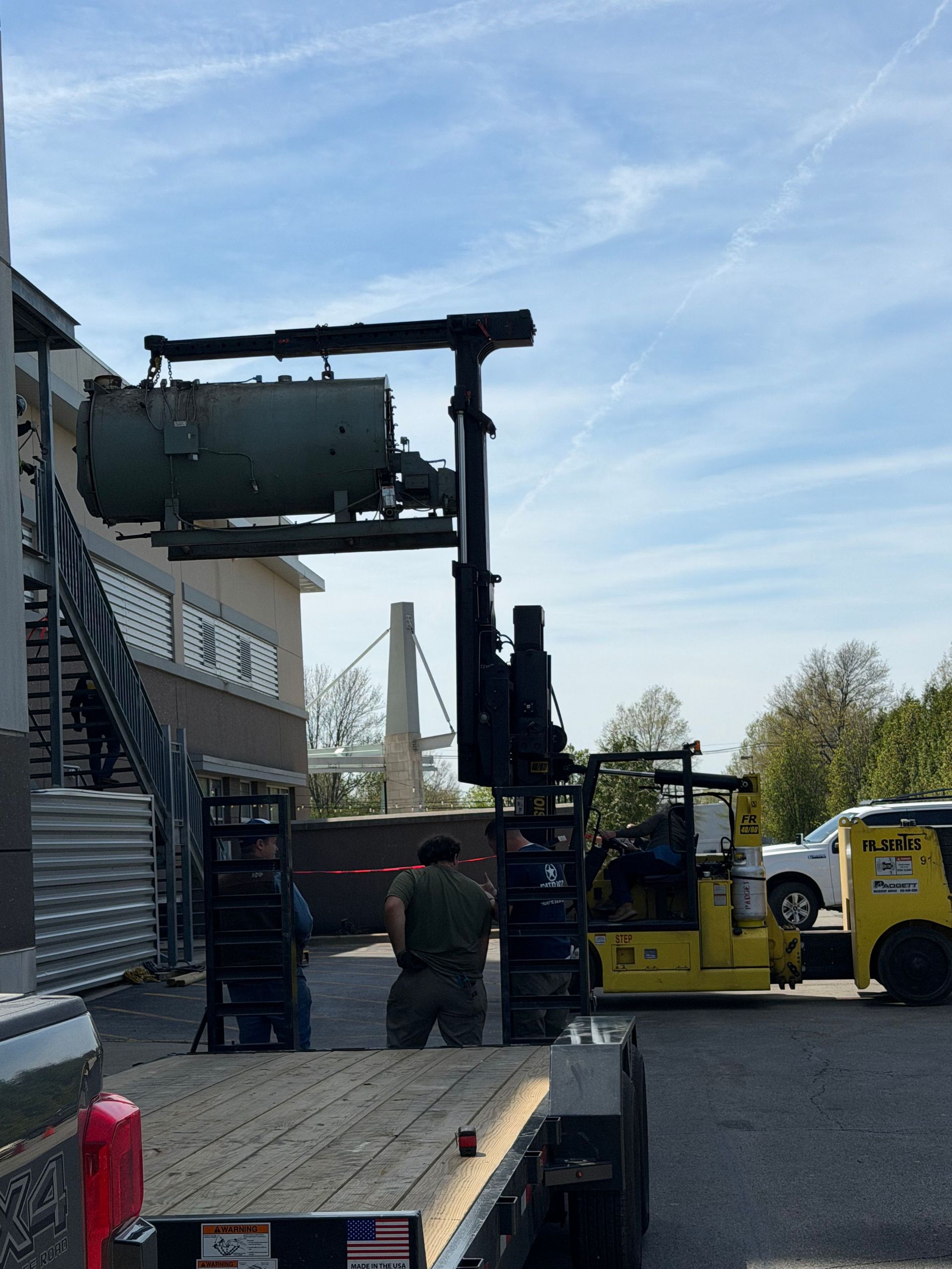 Forklift lifting a large metal tank beside a building, with workers on a paved lot. Forklift lifting a large metal tank beside a building, with workers on a paved lot.