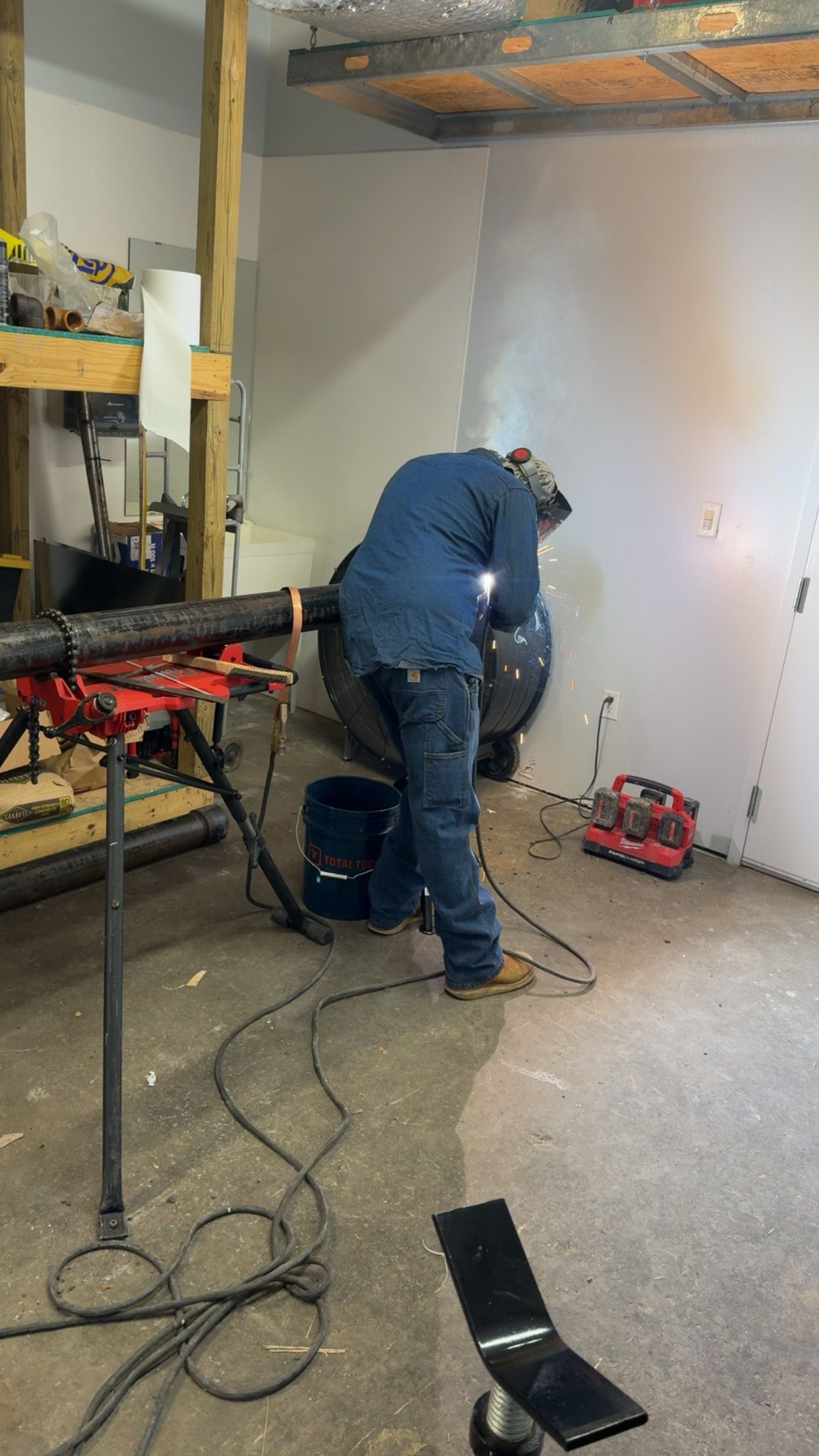 Worker using power tools in an unfinished room with exposed studs and concrete floor.
