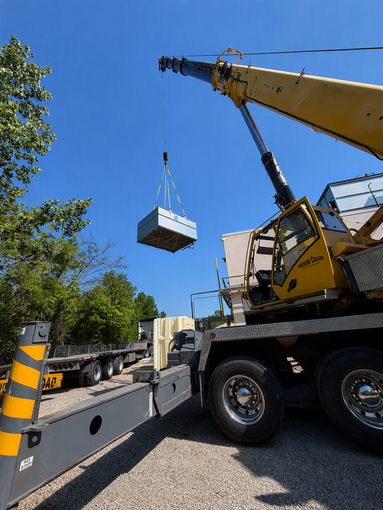 Crane lifting a gray box onto a flatbed truck beside a road under a blue sky