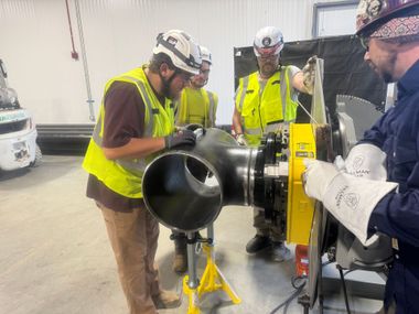 Workers in safety vests inspecting a large black pipe fitting in an industrial workshop.
