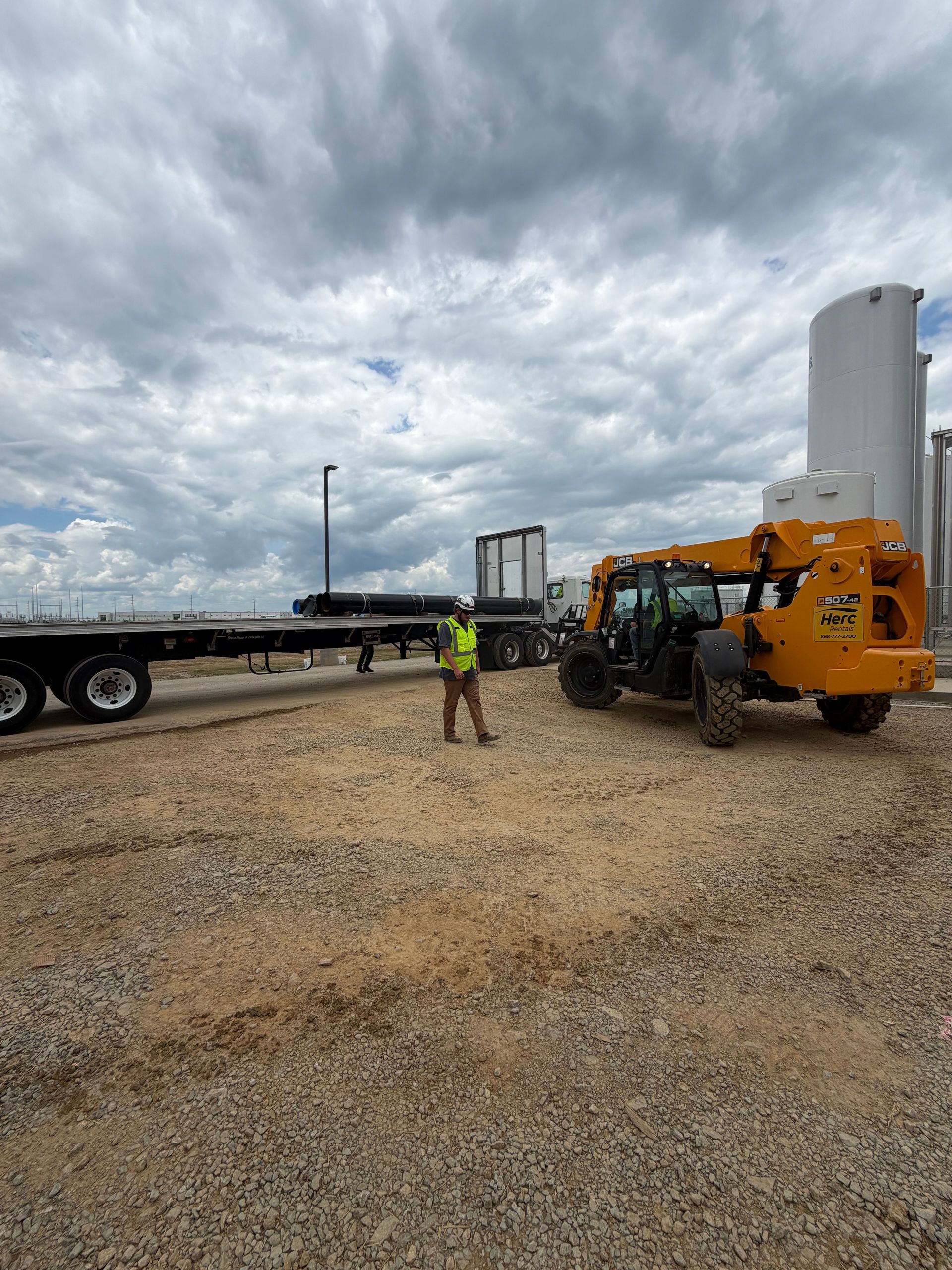 Worker stands beside orange heavy equipment on a gravel lot under a cloudy sky.