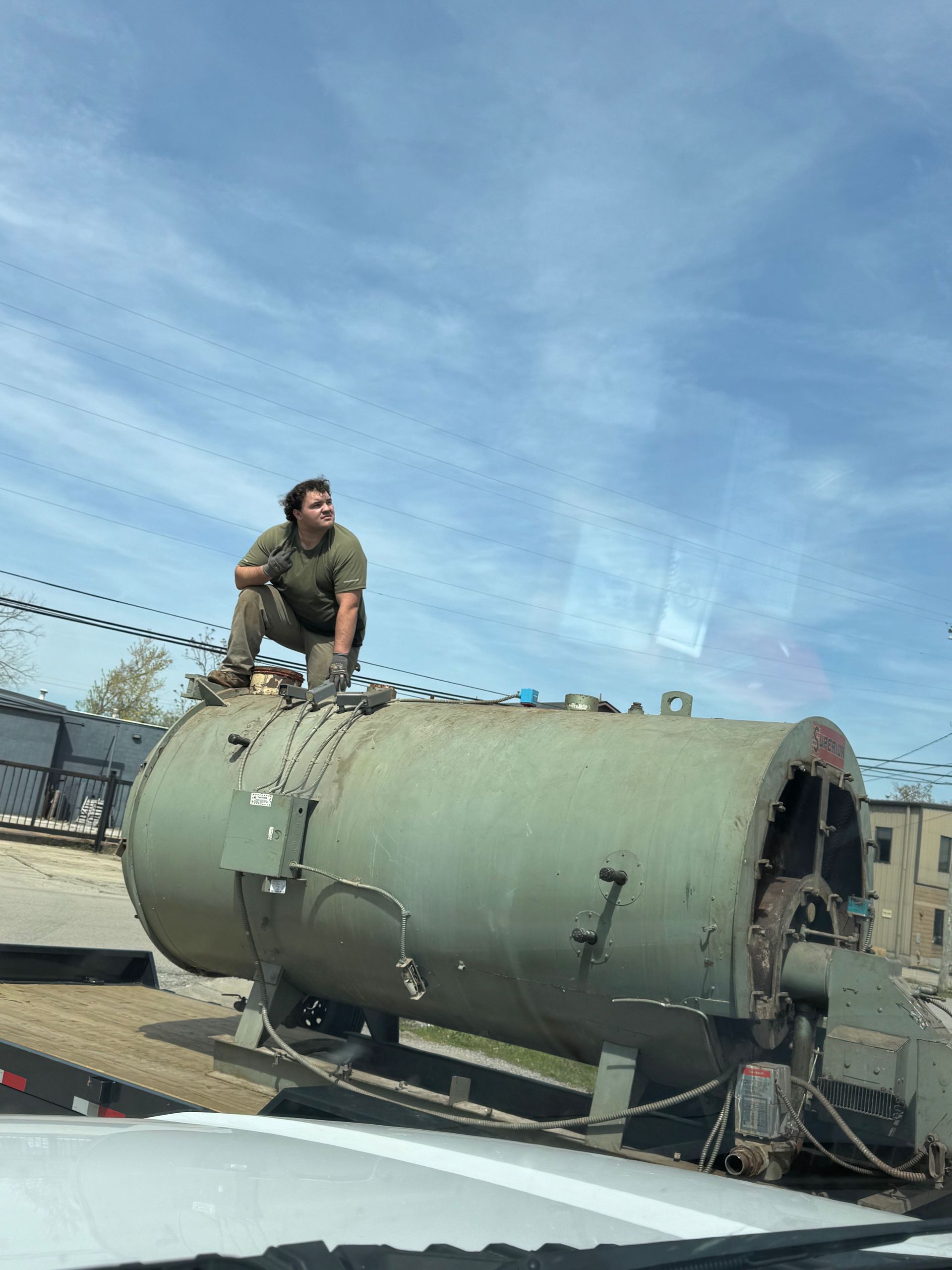 Worker crouches on a large green industrial tank outdoors under a blue sky.