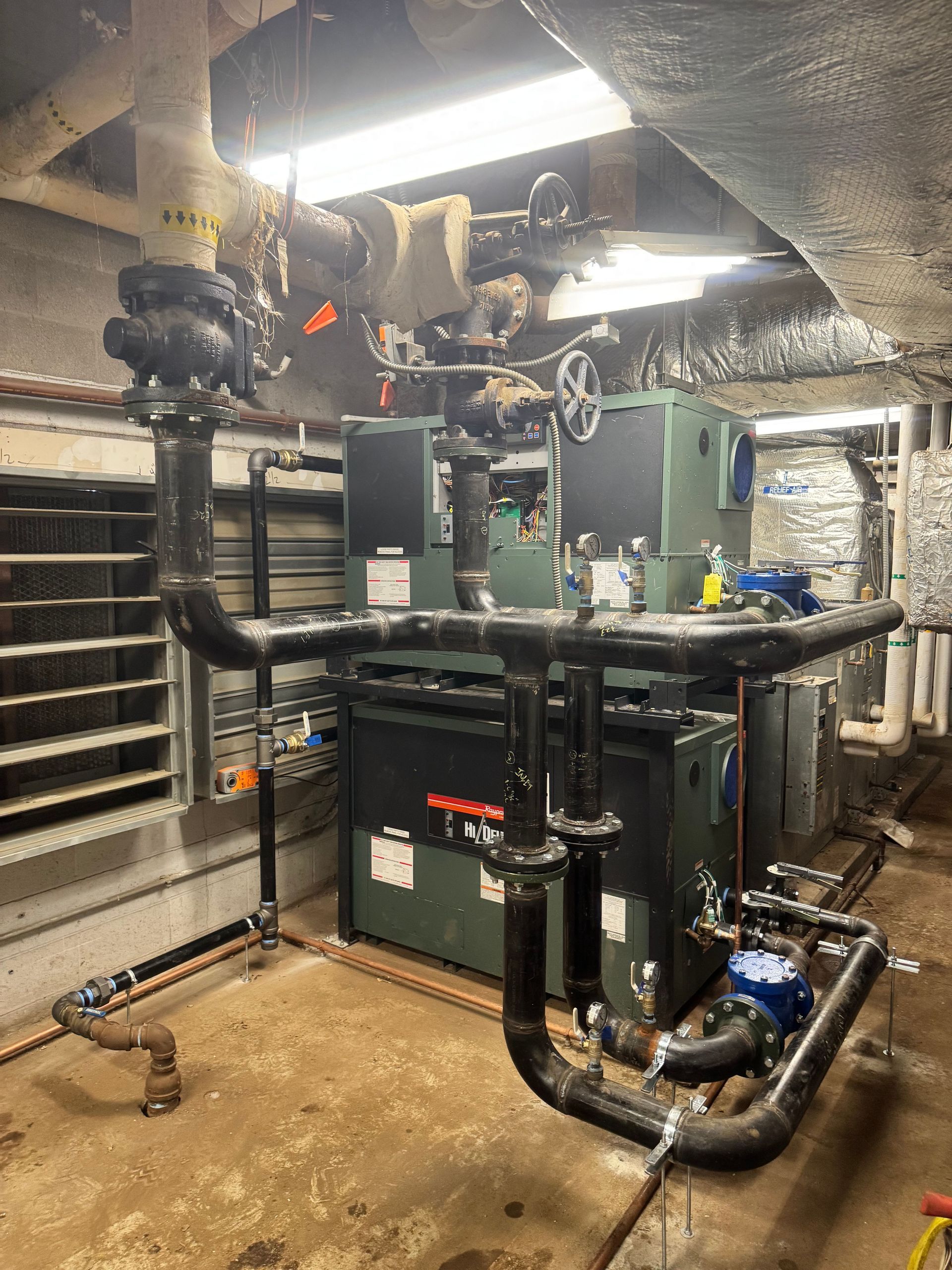 Industrial HVAC equipment in a basement room with large pipes, ducts, and machinery units.