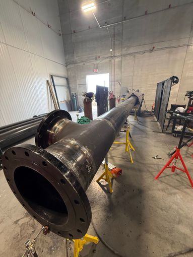 Large black industrial pipe assembly in a workshop, supported by stands in a bright metal-fabrication room