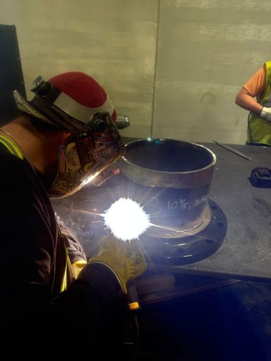Worker in a face shield welding a large metal cylinder indoors, with bright sparks visible.