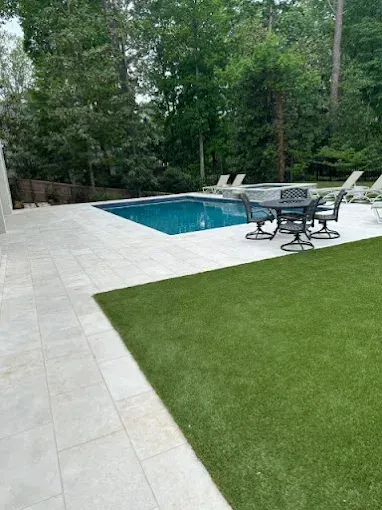 Pool area with light stone patio, green lawn, pool, and patio furniture.