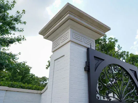 White brick pillar with decorative top and black gate, set outdoors with greenery and sky visible.